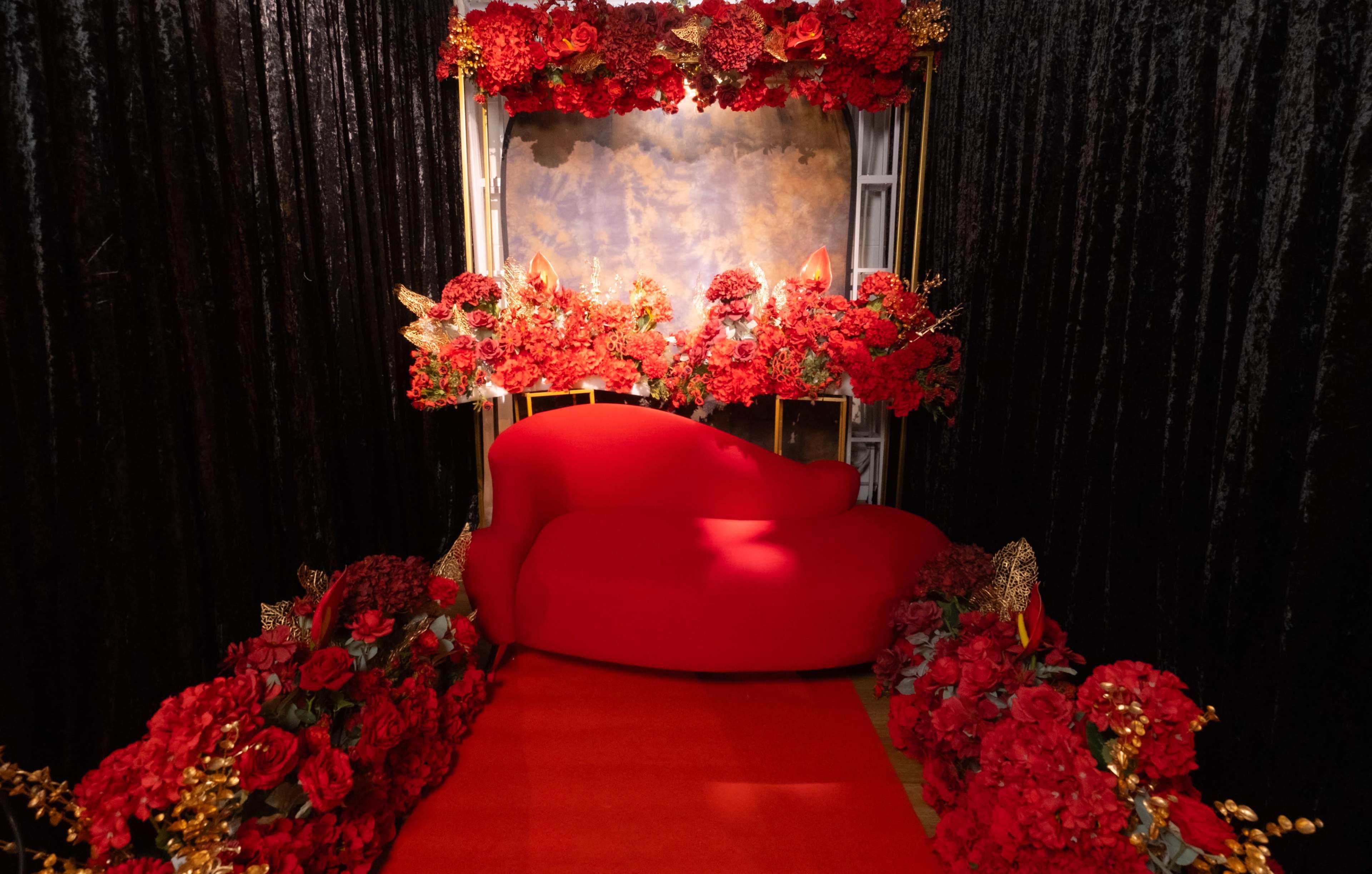 A red sofa surrounded by an arrangement of red flowers and a black backdrop, creating a vibrant display with a red carpet leading up to it.