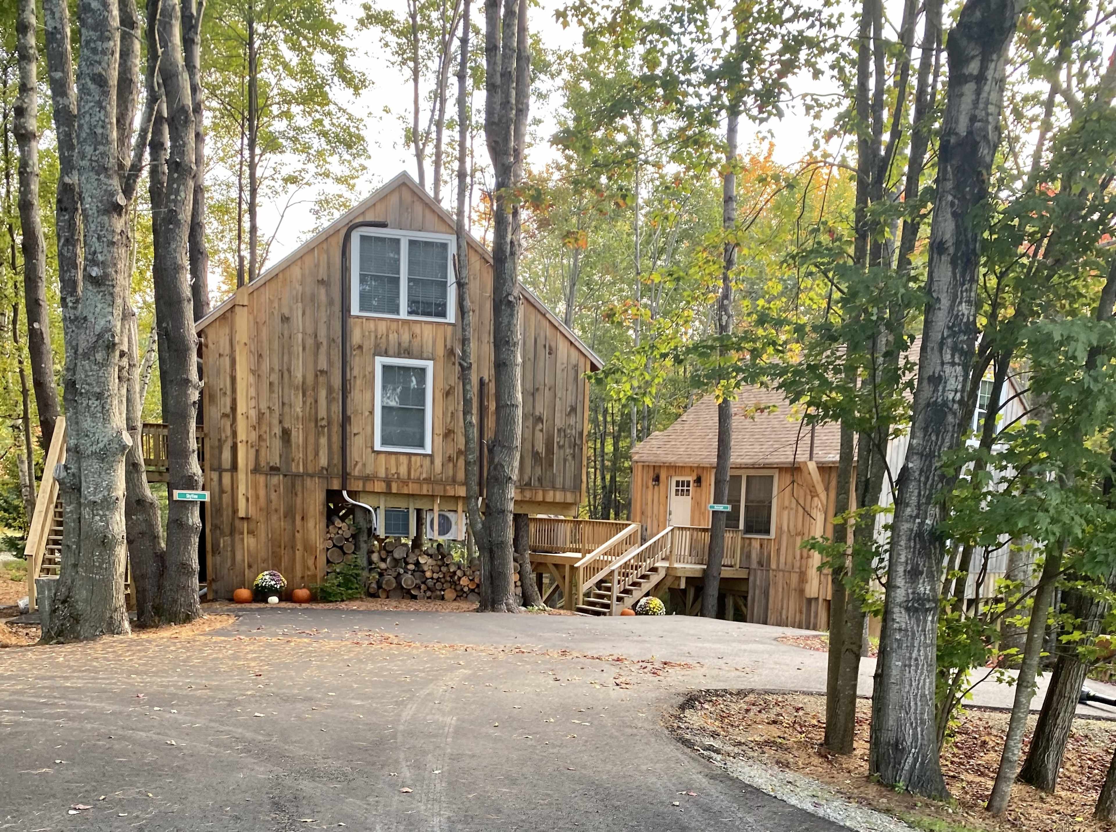 A wooden house with large windows sits among trees, featuring a driveway that curves around to the entrance.