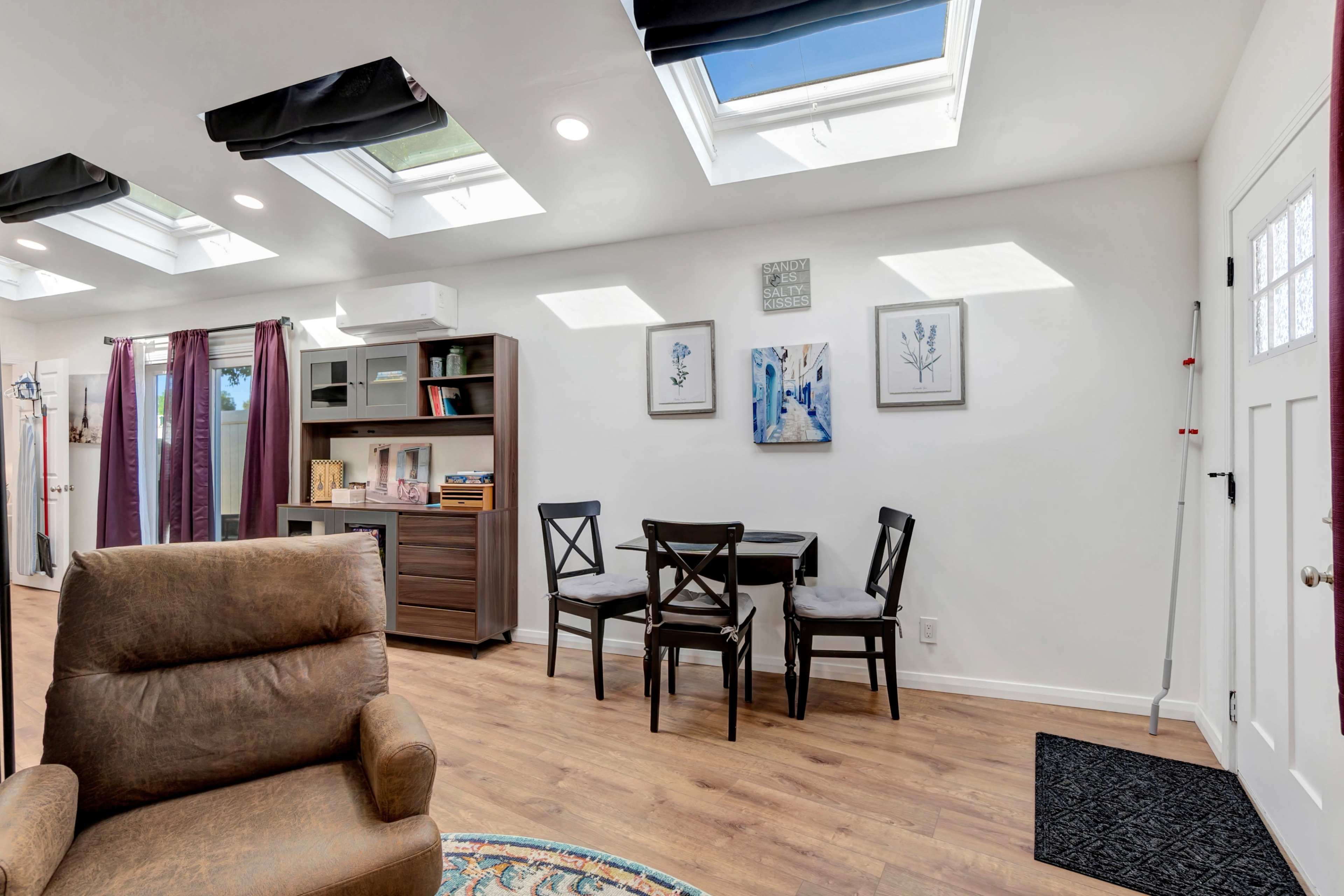 The image shows a bright room with skylights, a small dining table with four chairs, a brown armchair, and various decorative items on the walls.