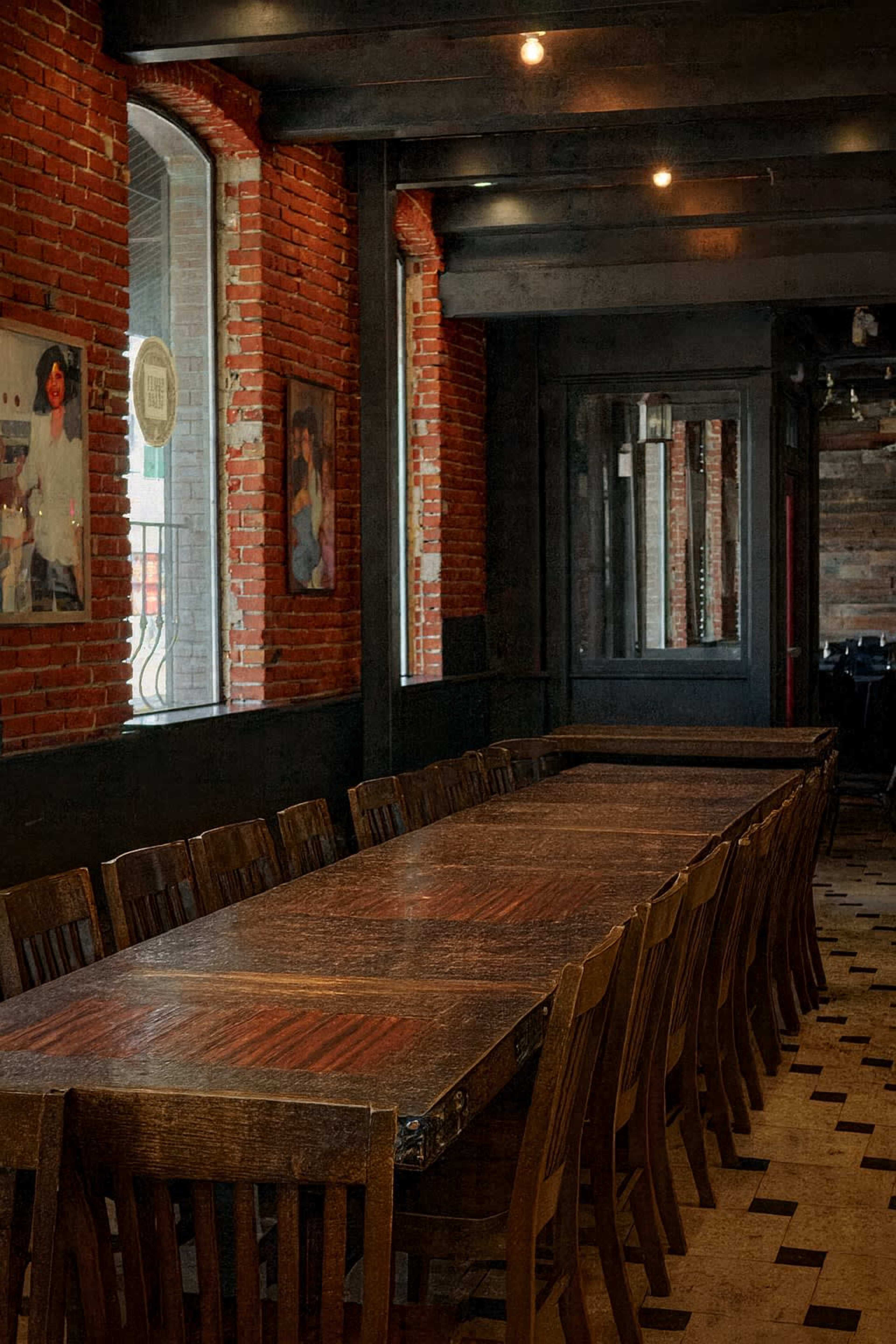 The image shows a large wooden dining table surrounded by wooden chairs in a brick-walled room with framed pictures on the walls.