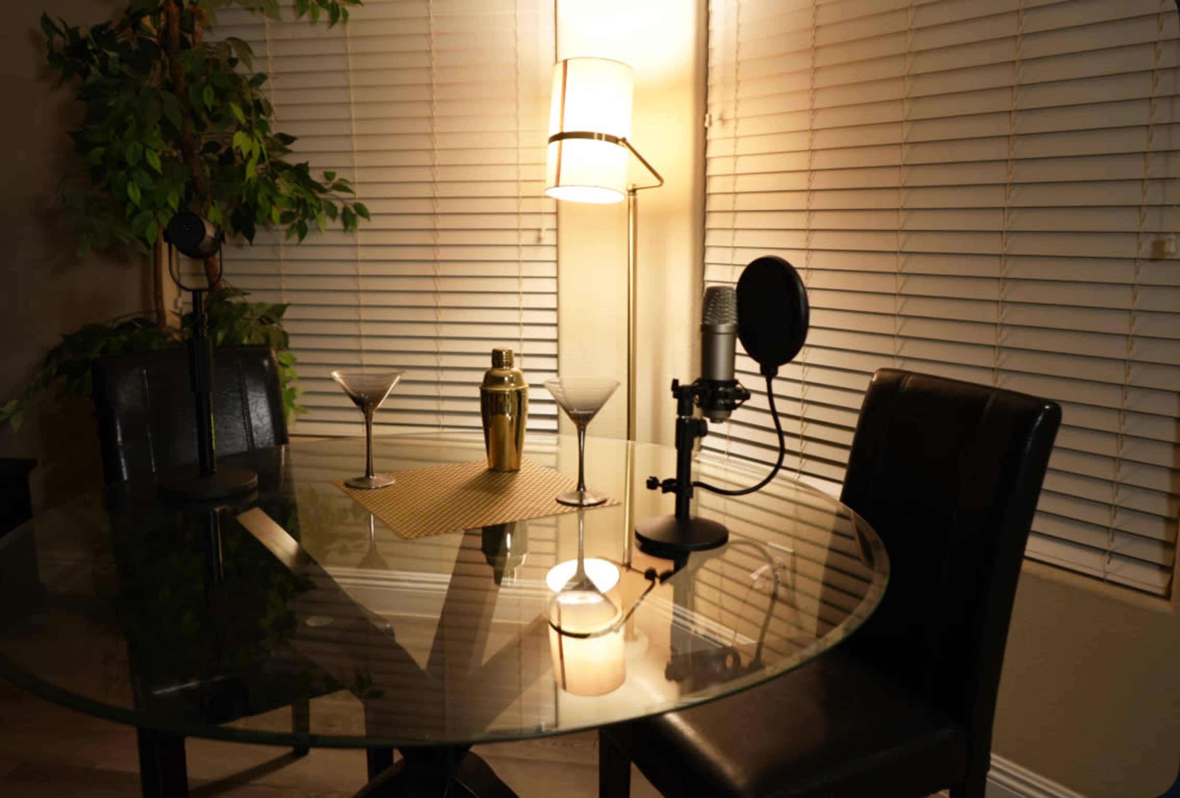 A round glass table is set with two martini glasses, a cocktail shaker, and microphone stands under a lamp next to a potted plant.