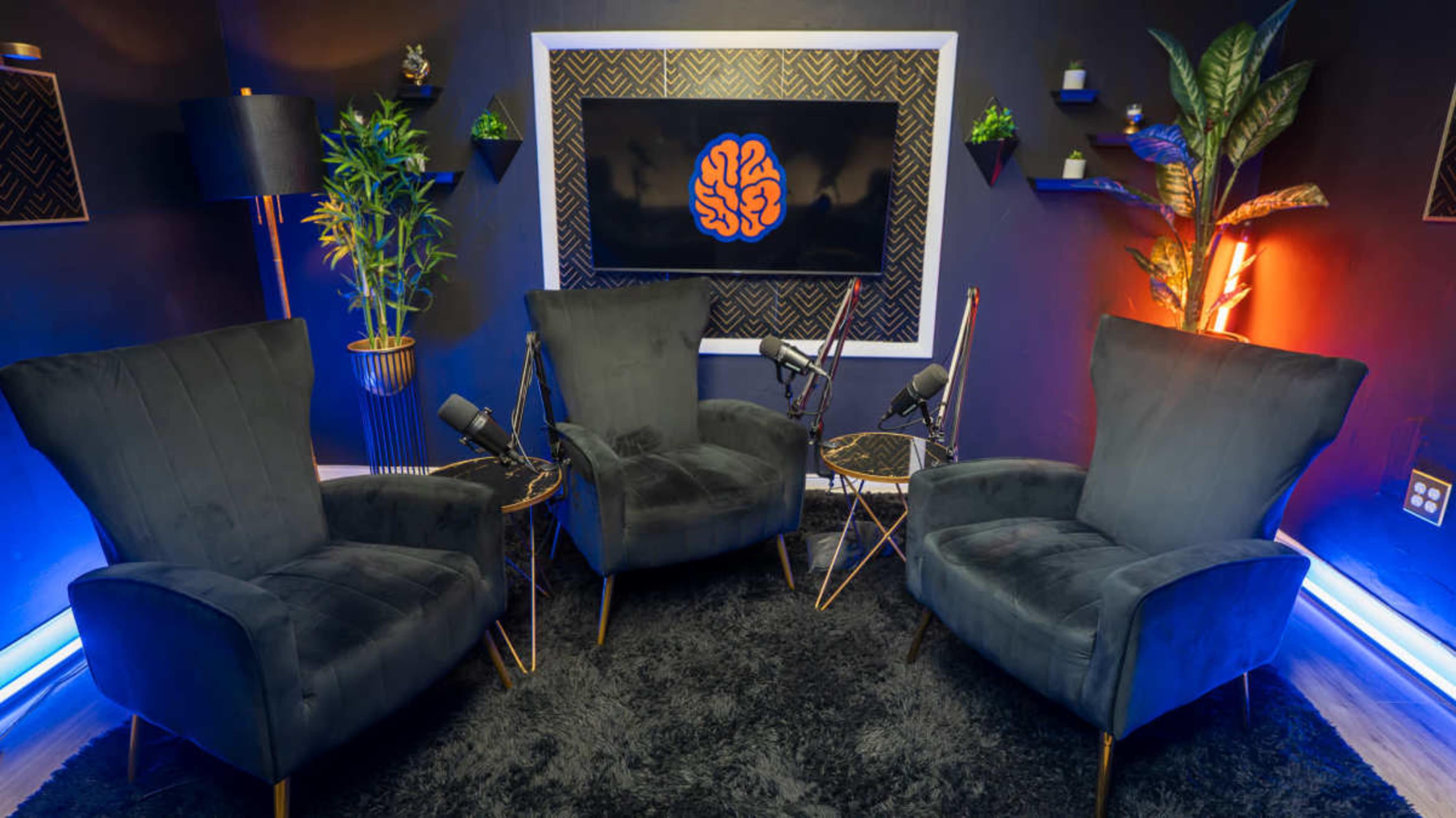 The image shows a cozy podcast studio setup featuring three velvet chairs, a large wall-mounted screen with a brain graphic, and various plants and lighting.