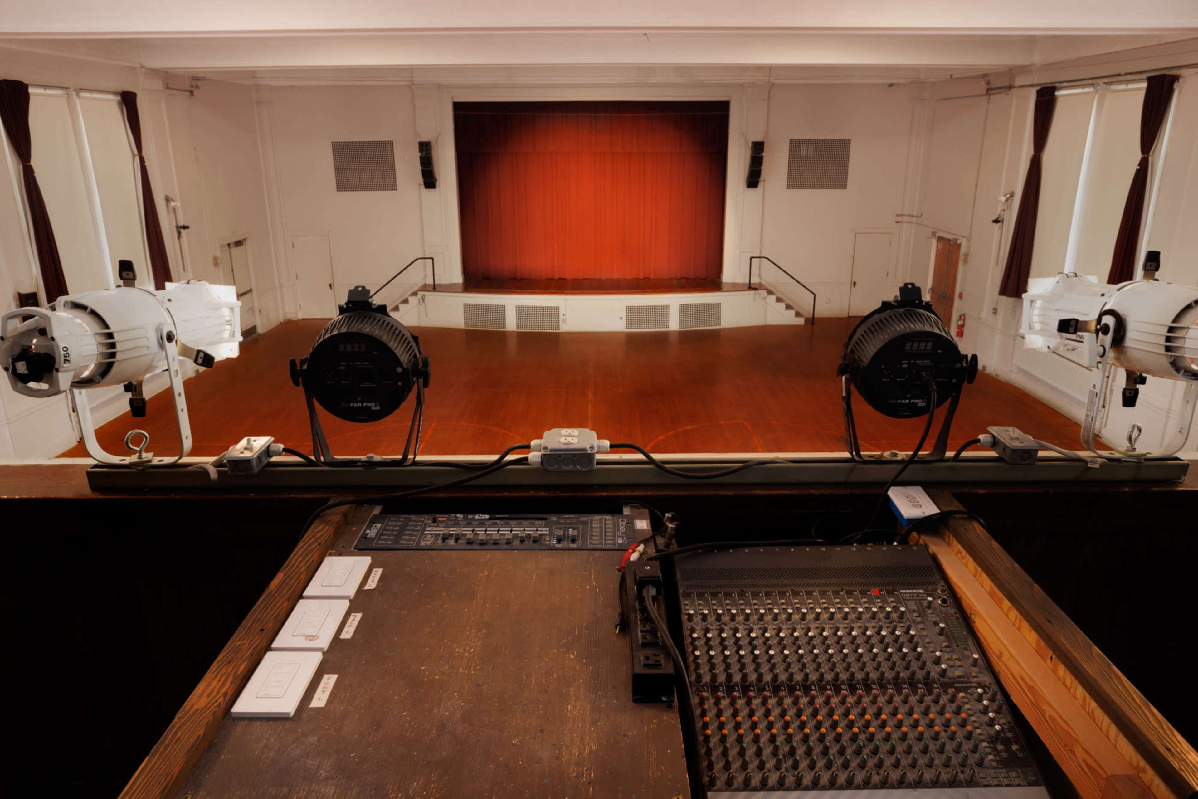 A well-lit theater stage with a red curtain and a sound mixing console in the foreground.