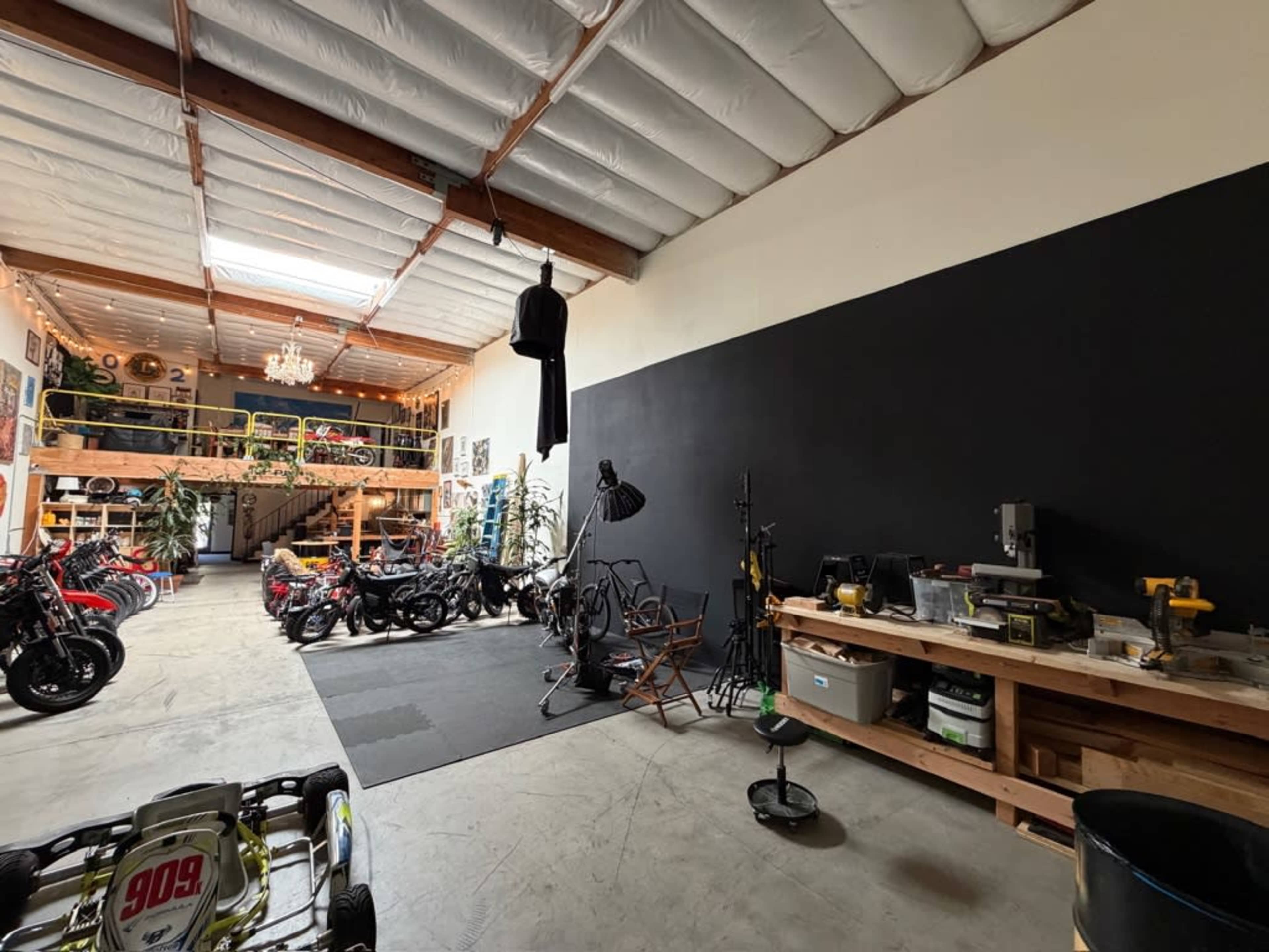 The image shows a spacious workshop or garage with a black wall and various motorcycles, tools, and workstations arranged throughout the area.