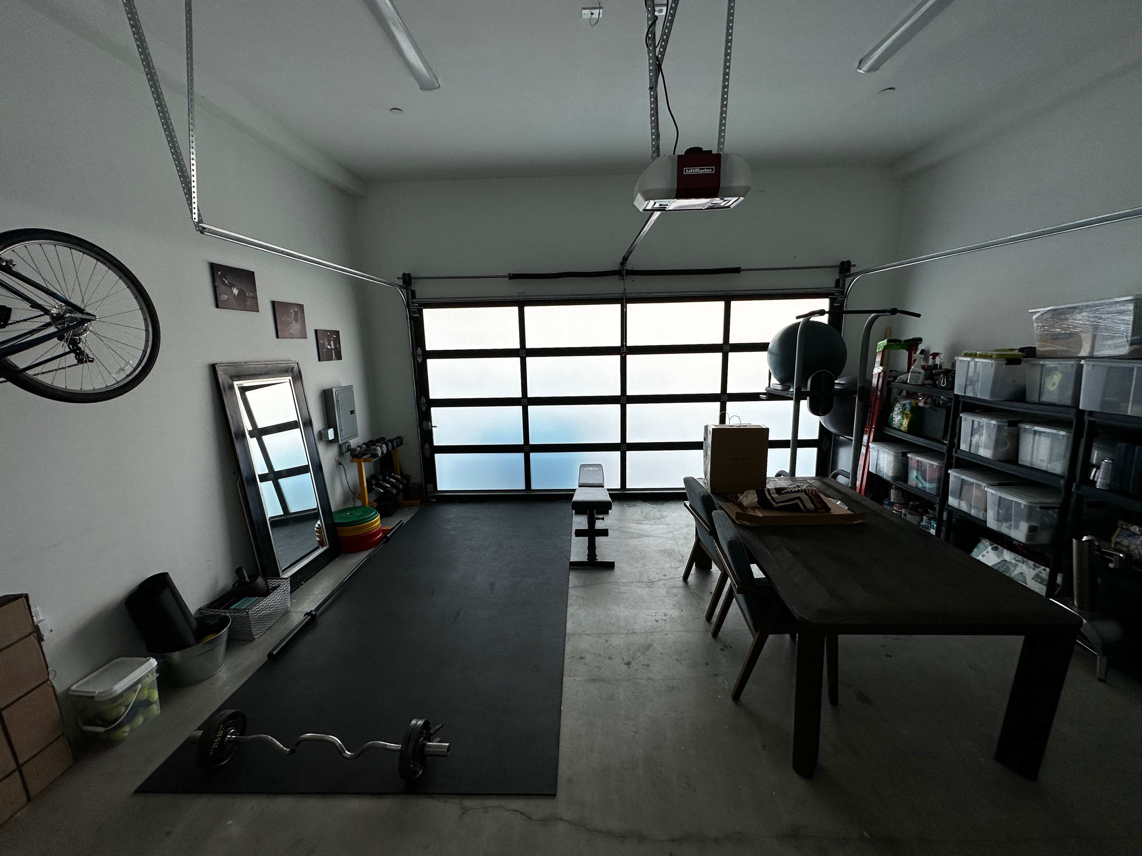 A garage space features workout equipment, a table, storage shelves, a mirror, and a bicycle hung on the wall.