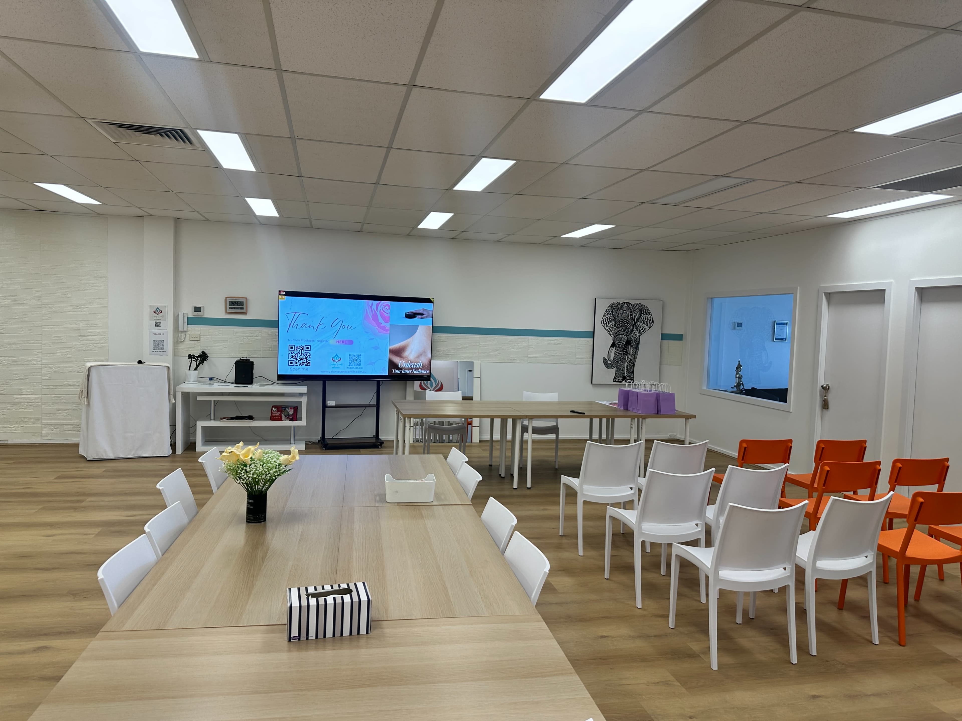 The image shows a well-lit meeting room with a large screen at the front, white chairs arranged around a wooden table, and a few decorative elements on the table.