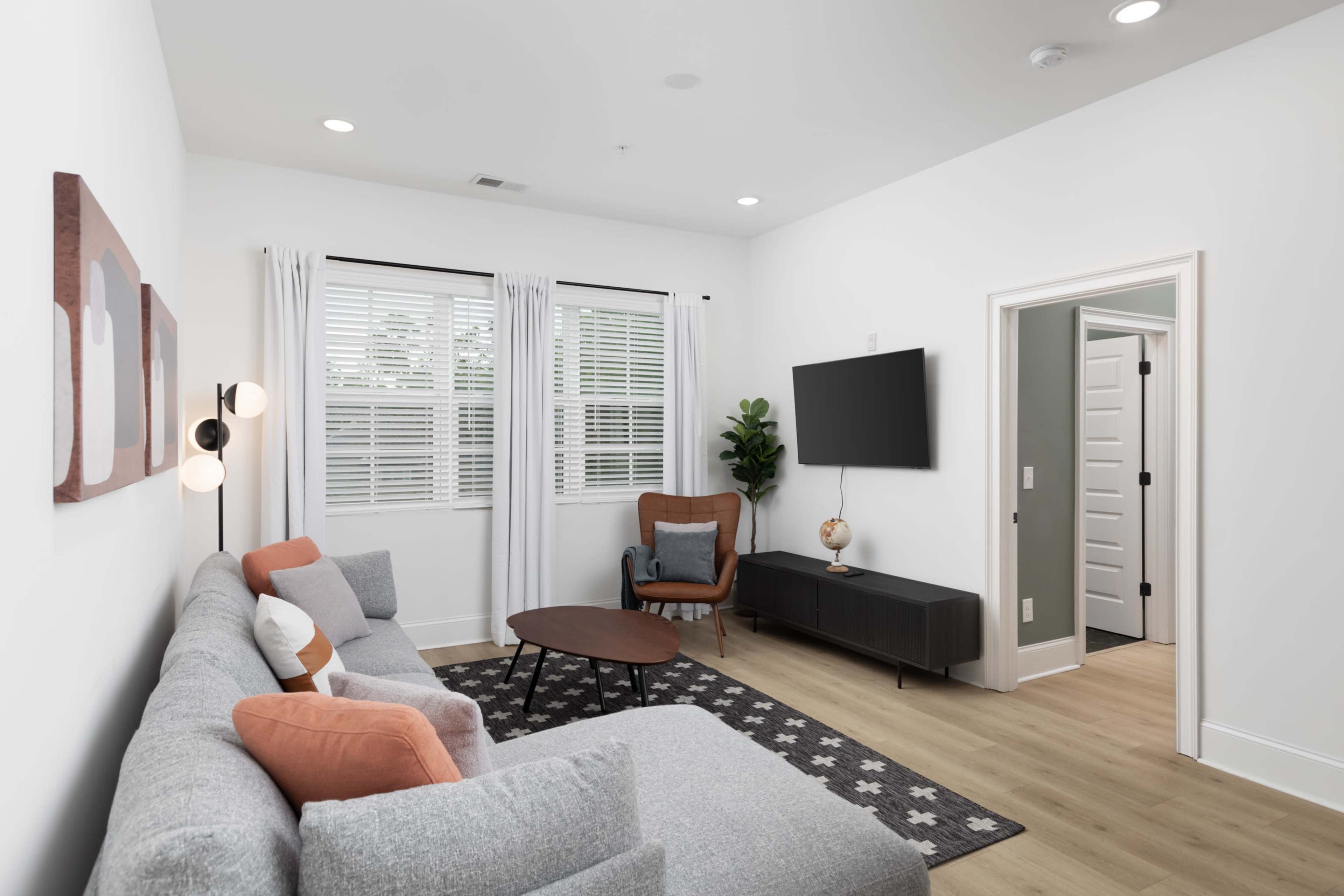 A modern living room features a gray sectional sofa, a wooden coffee table, a wall-mounted TV, and a chair beside a window.