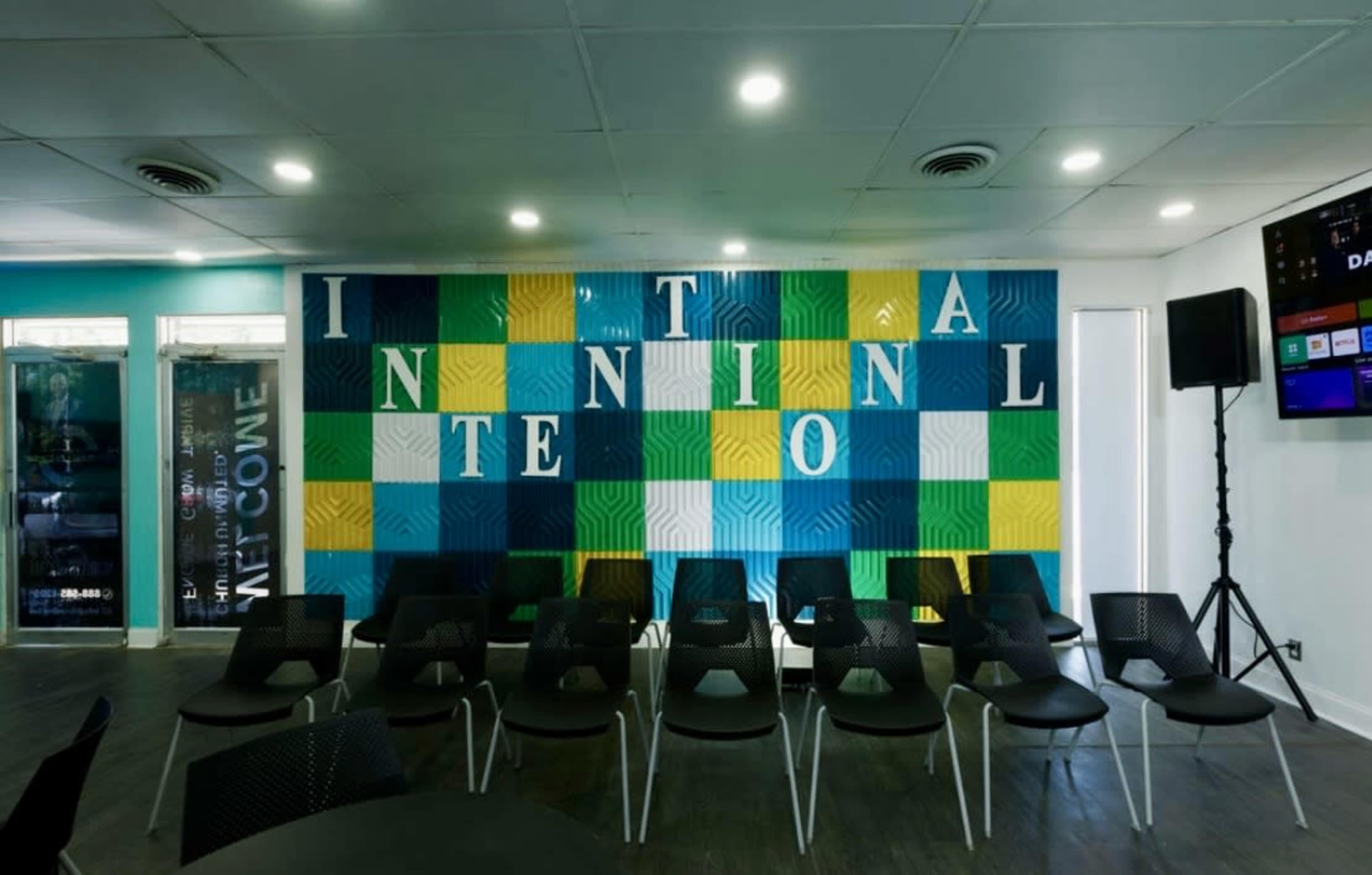 The image shows a colorful wall with the word "INTENTIONAL" displayed in large letters, alongside rows of black chairs arranged for seating in a modern indoor space.