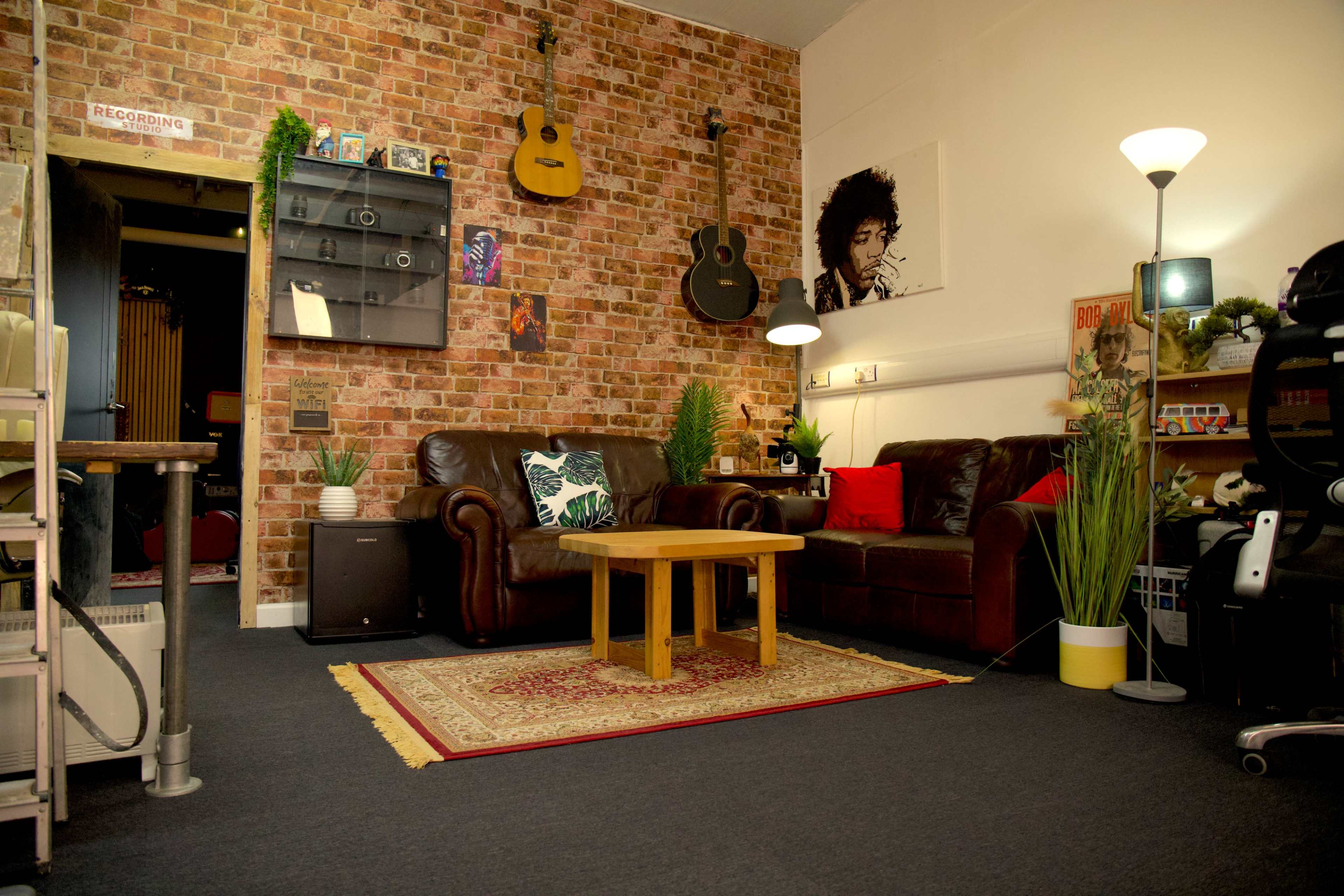 The image features a cozy room with a brick-patterned wall, two leather sofas, a wooden coffee table, and various musical instruments and decorations, including a portrait on the wall.