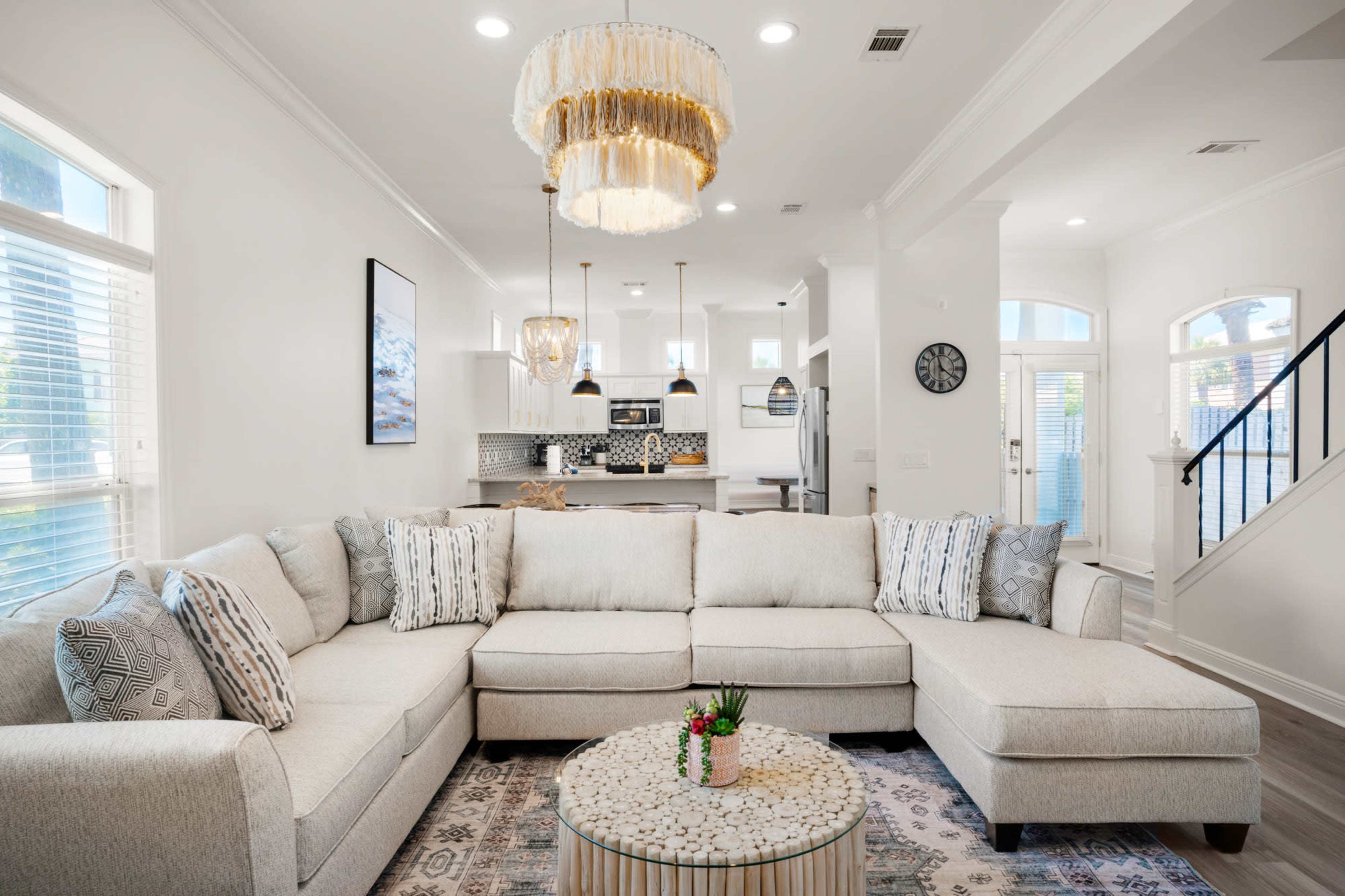 A modern living room features a large sectional sofa, a round coffee table, and a chandelier, with a view of an open kitchen in the background.