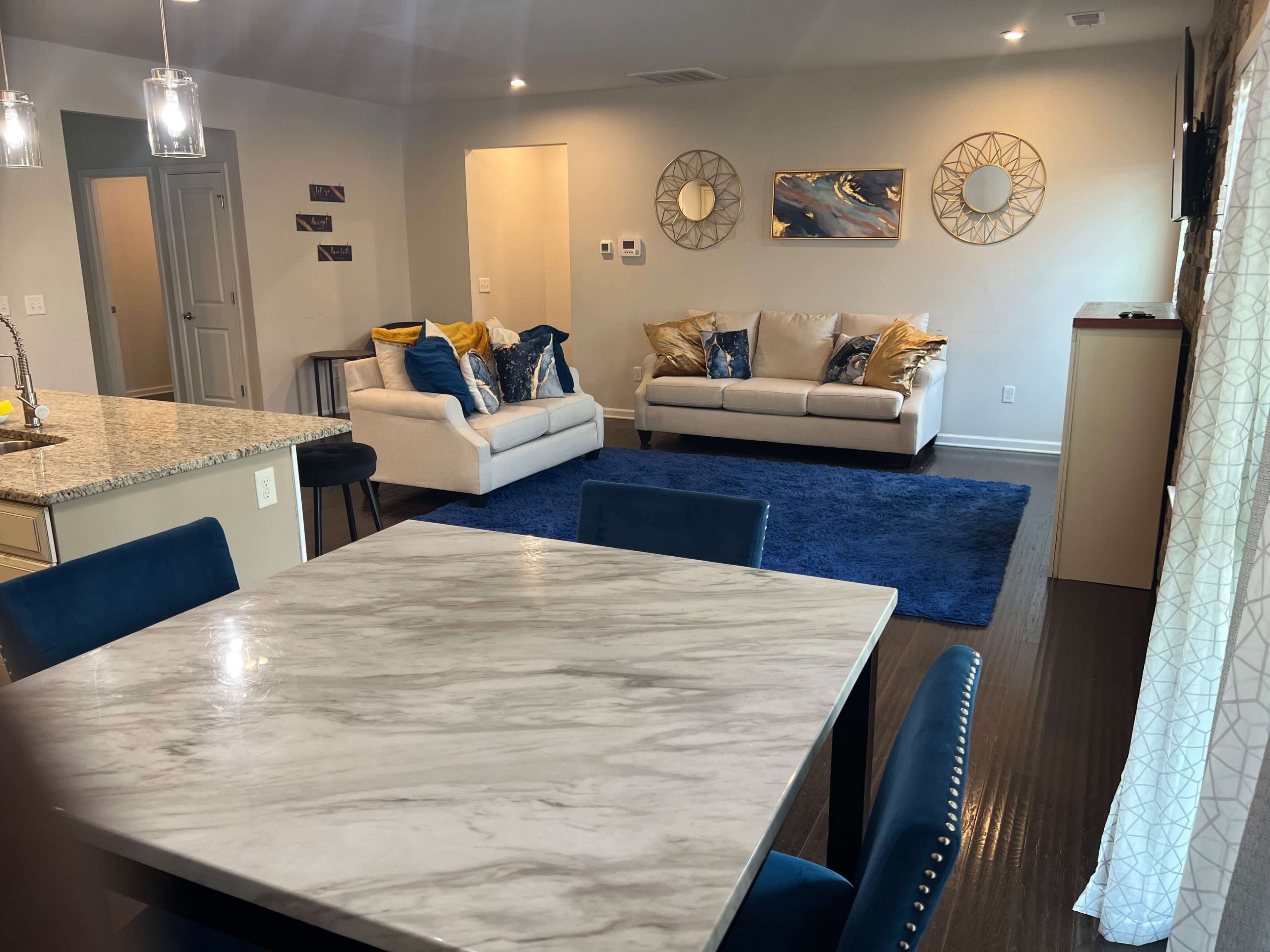 A modern living room and dining area featuring a marble dining table, blue upholstered chairs, two sofas with decorative pillows, and a blue rug on a wooden floor.