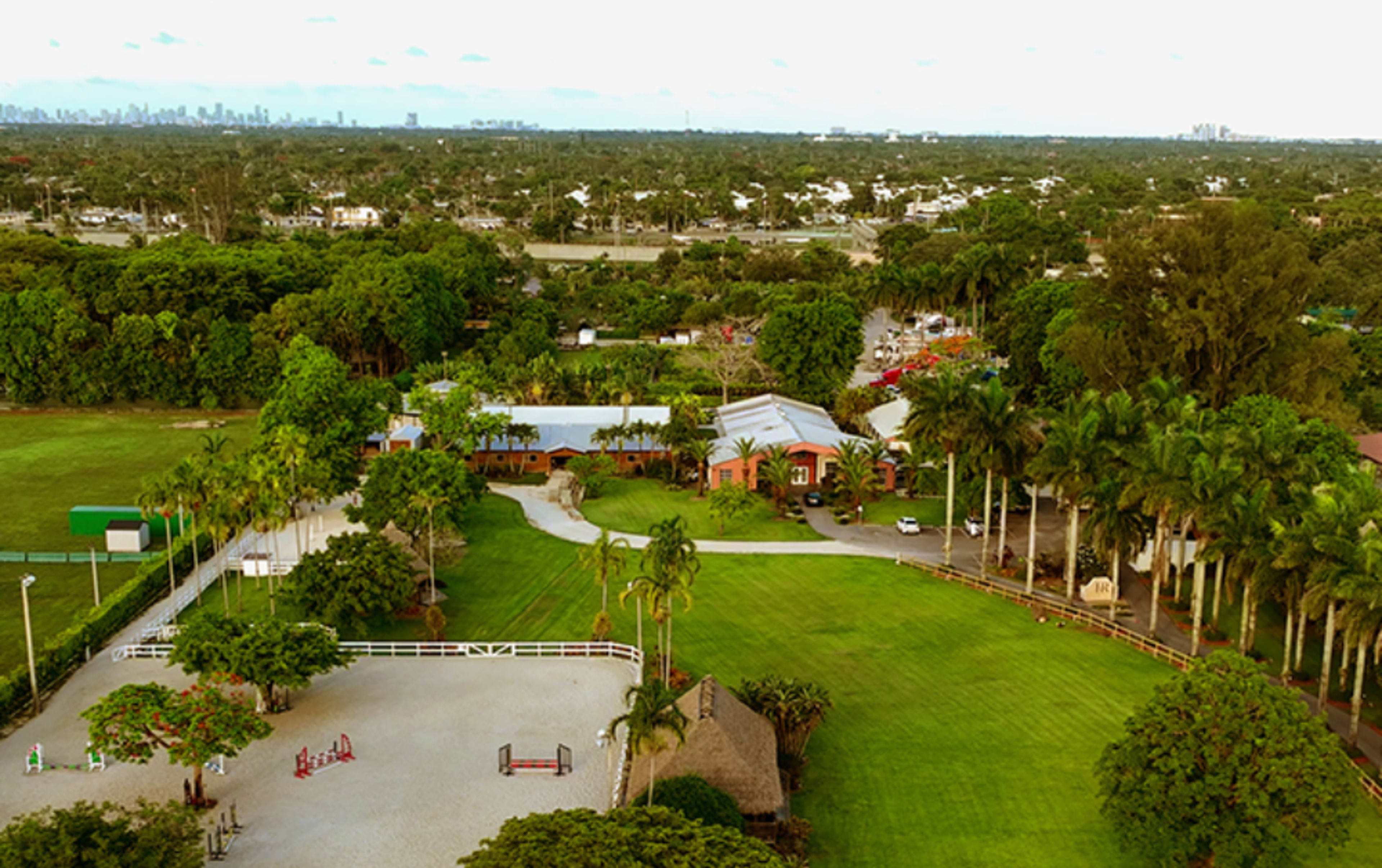 The image shows an expansive equestrian facility surrounded by lush greenery, with a clear view of a city skyline in the background.