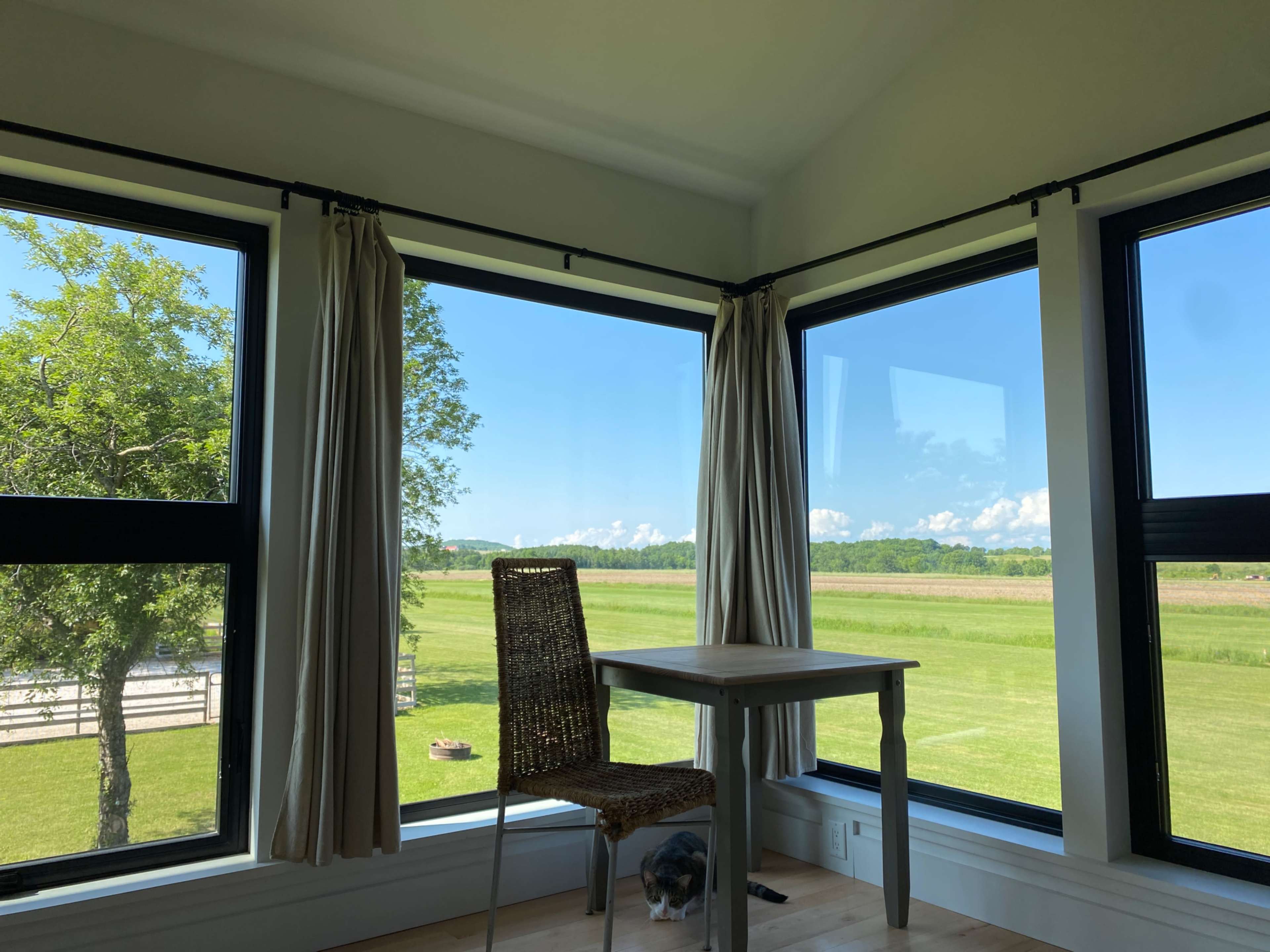A table and chair are positioned near large windows that offer a view of a grassy field and a blue sky.