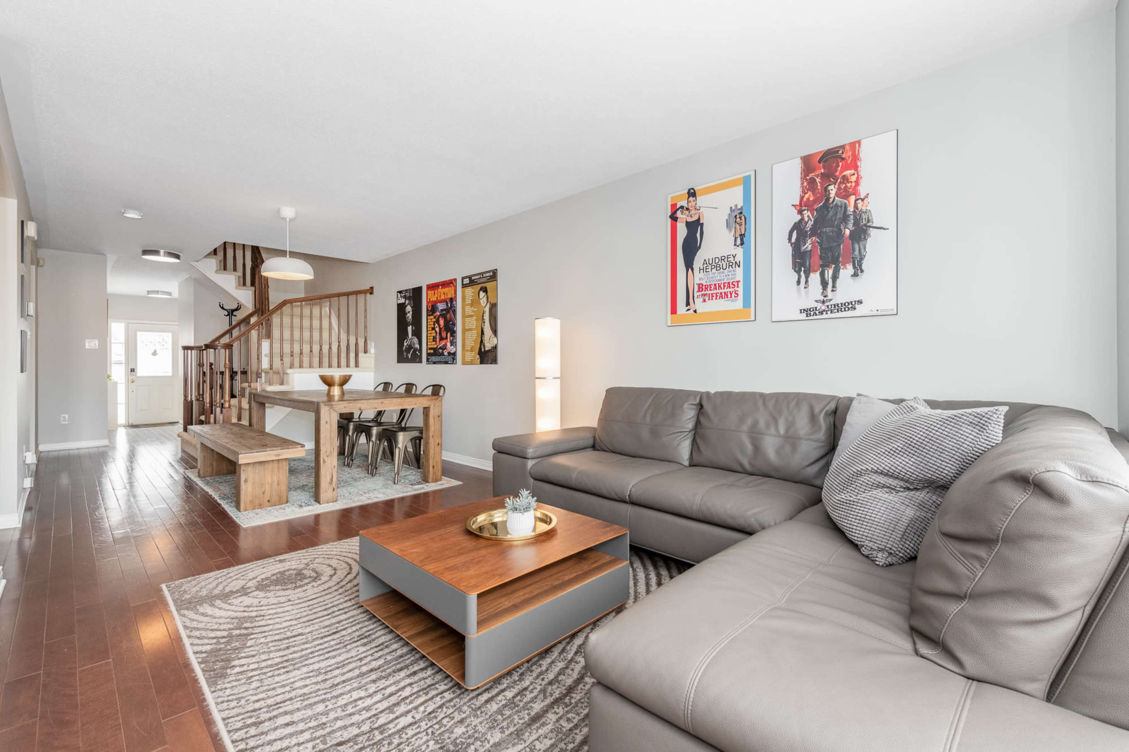 A modern living room with a gray sectional sofa, a wooden coffee table, and vintage movie posters on the walls, adjacent to a staircase leading to an upper level.