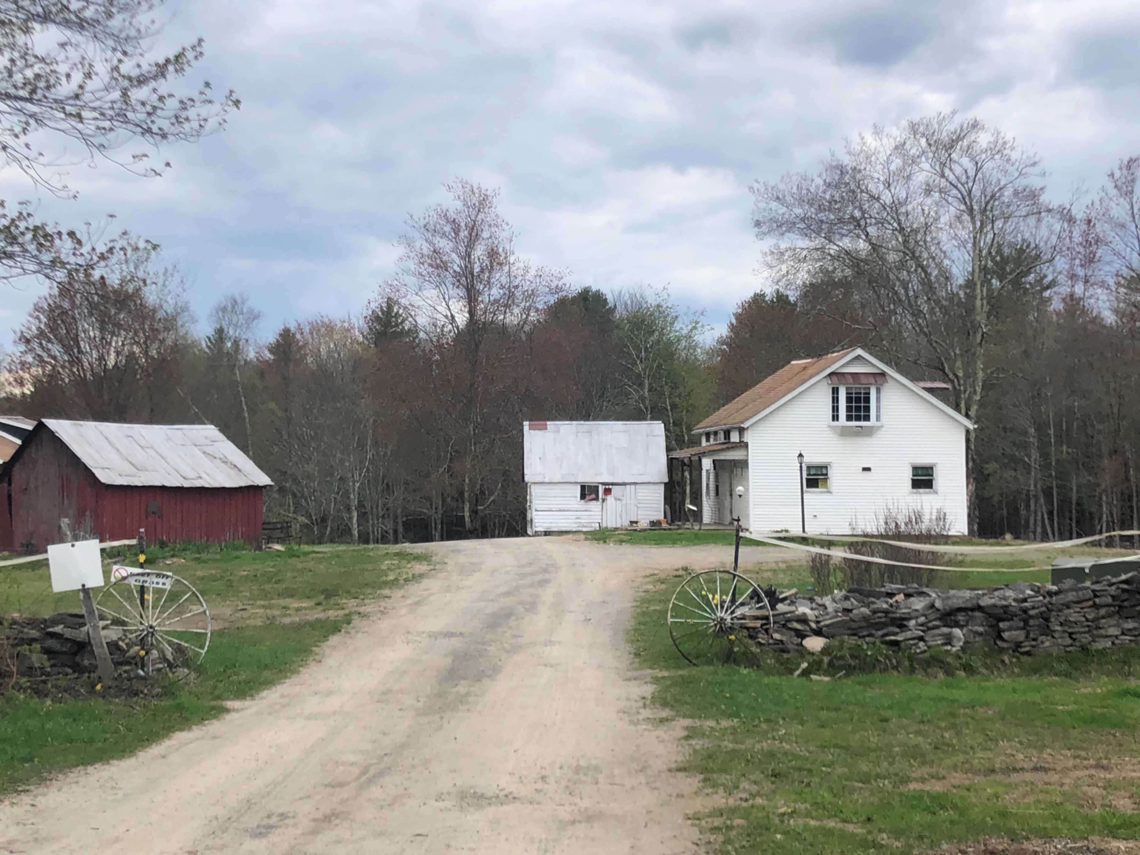 A dirt road leads past two structures, one red and one white, with trees in the background and a stone wall along the side.