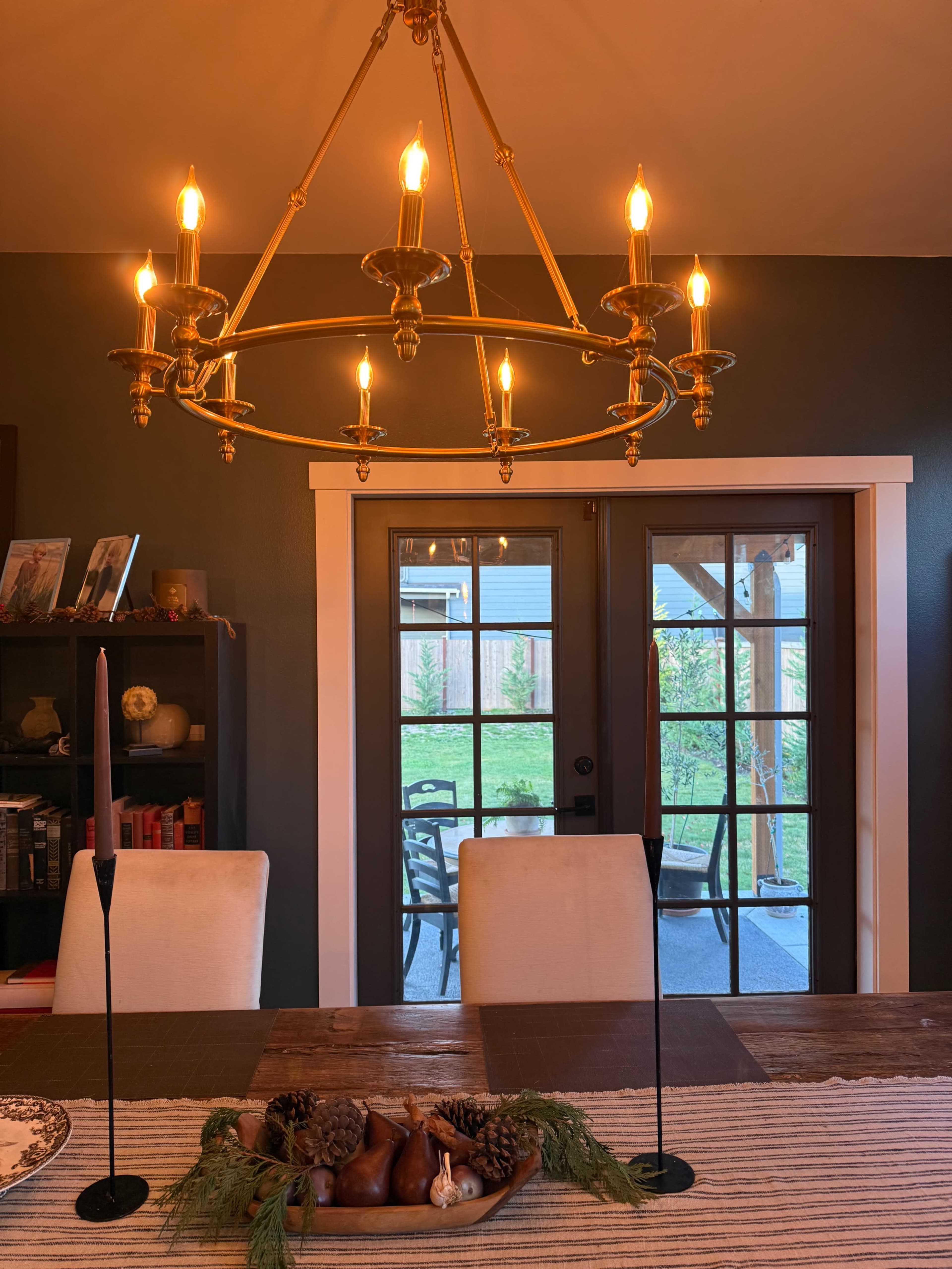 A decorative chandelier hangs above a dining table, which is set with candles and a centerpiece of pinecones and pears, while double doors lead to a patio area visible in the background.