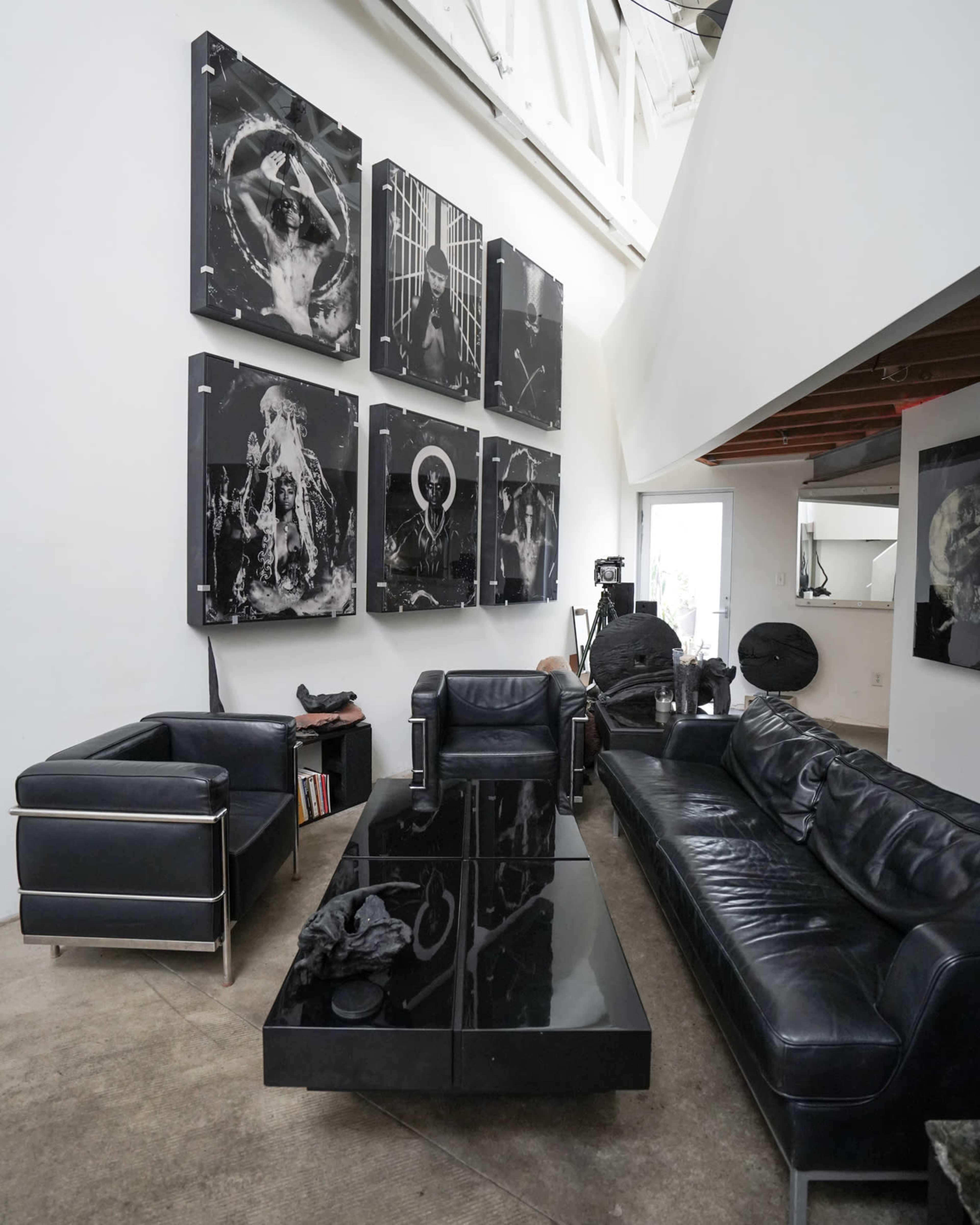 The image shows a modern living space featuring black leather couches, a glass coffee table, and several large black-and-white photographs on the wall.