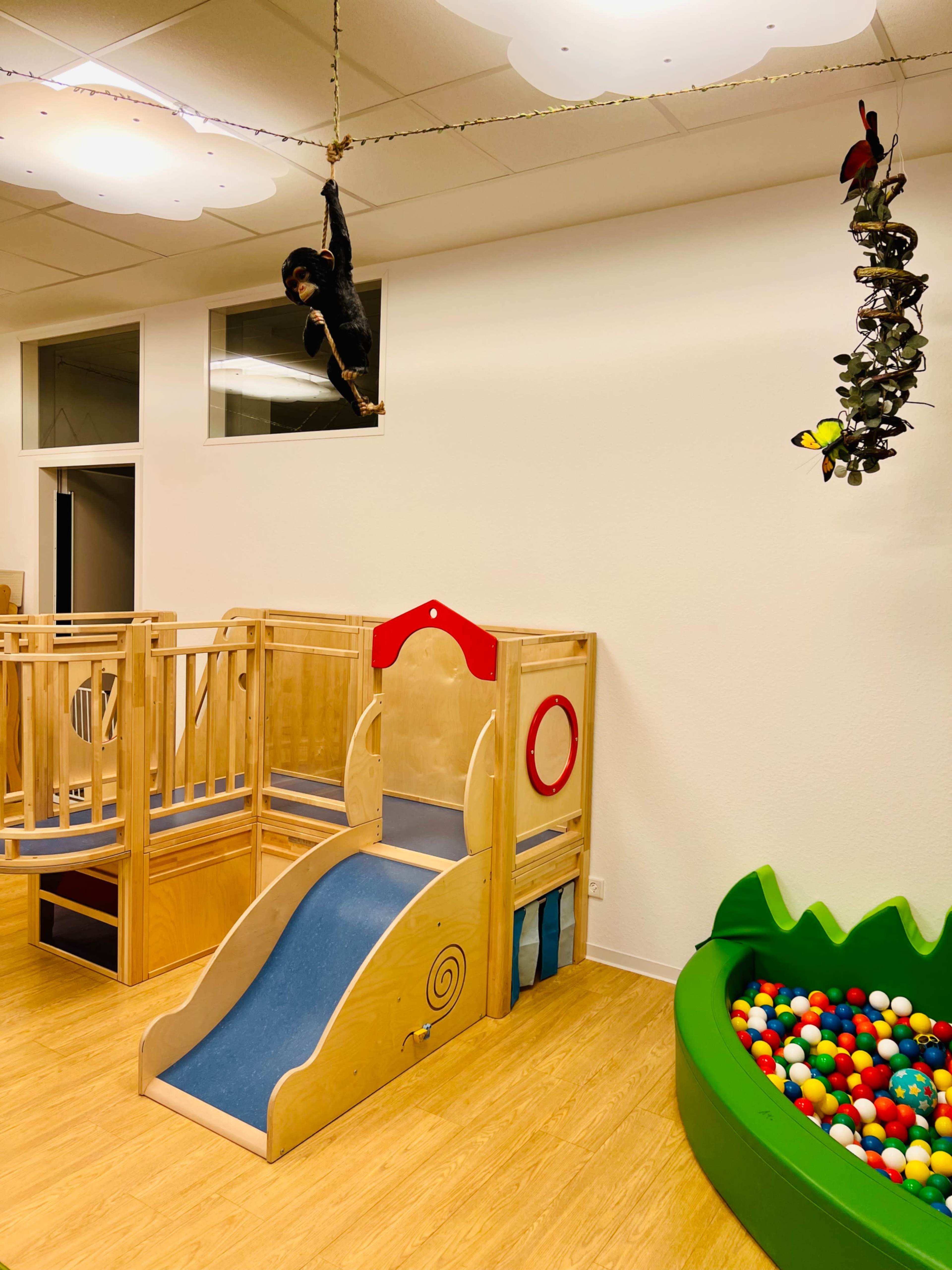 The image shows a brightly lit children's play area featuring a wooden slide, climbing structure, and a ball pit filled with colorful balls.