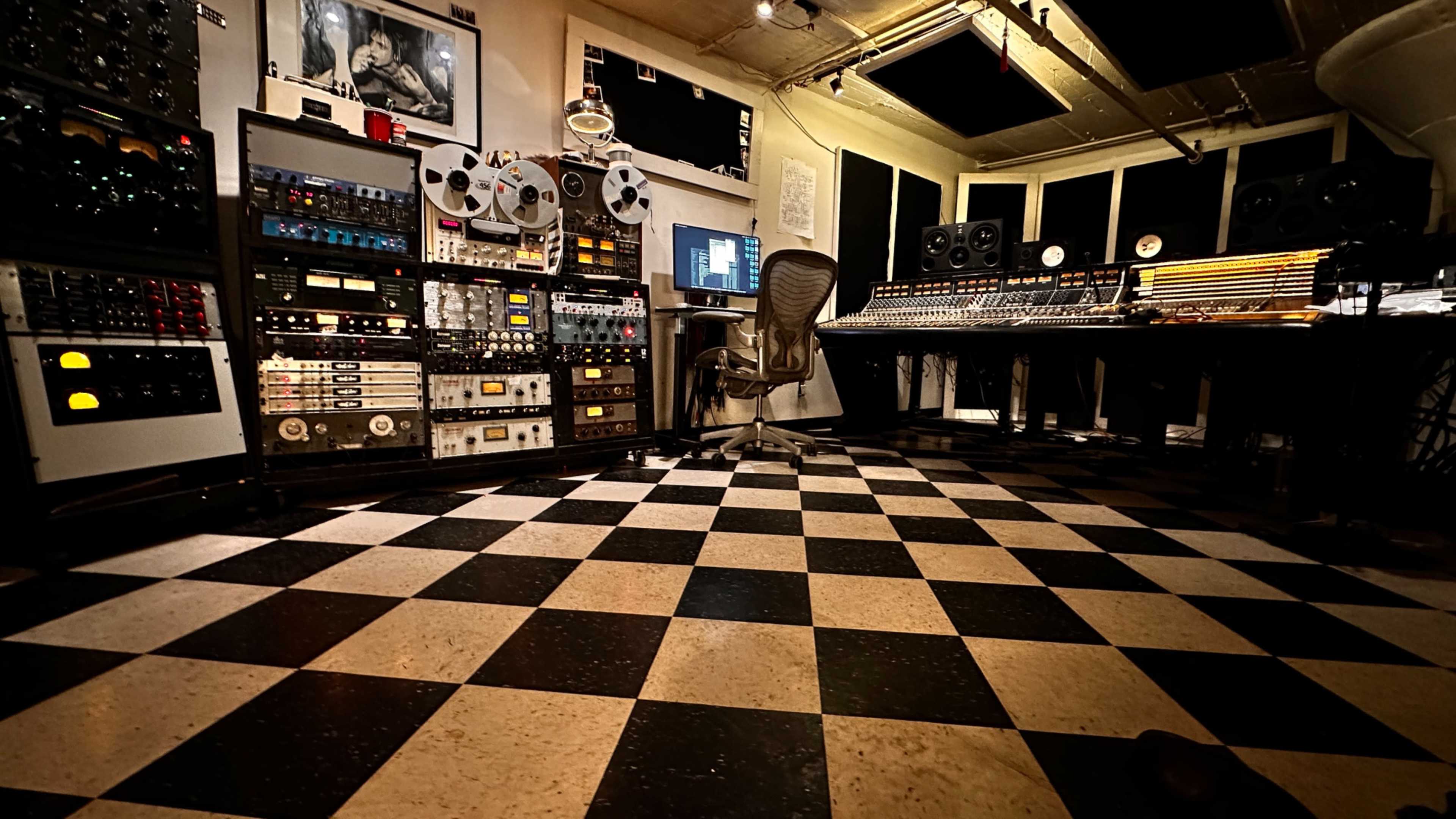 The image shows a recording studio with various audio equipment, including tape machines and mixing consoles, arranged around a checkered floor.