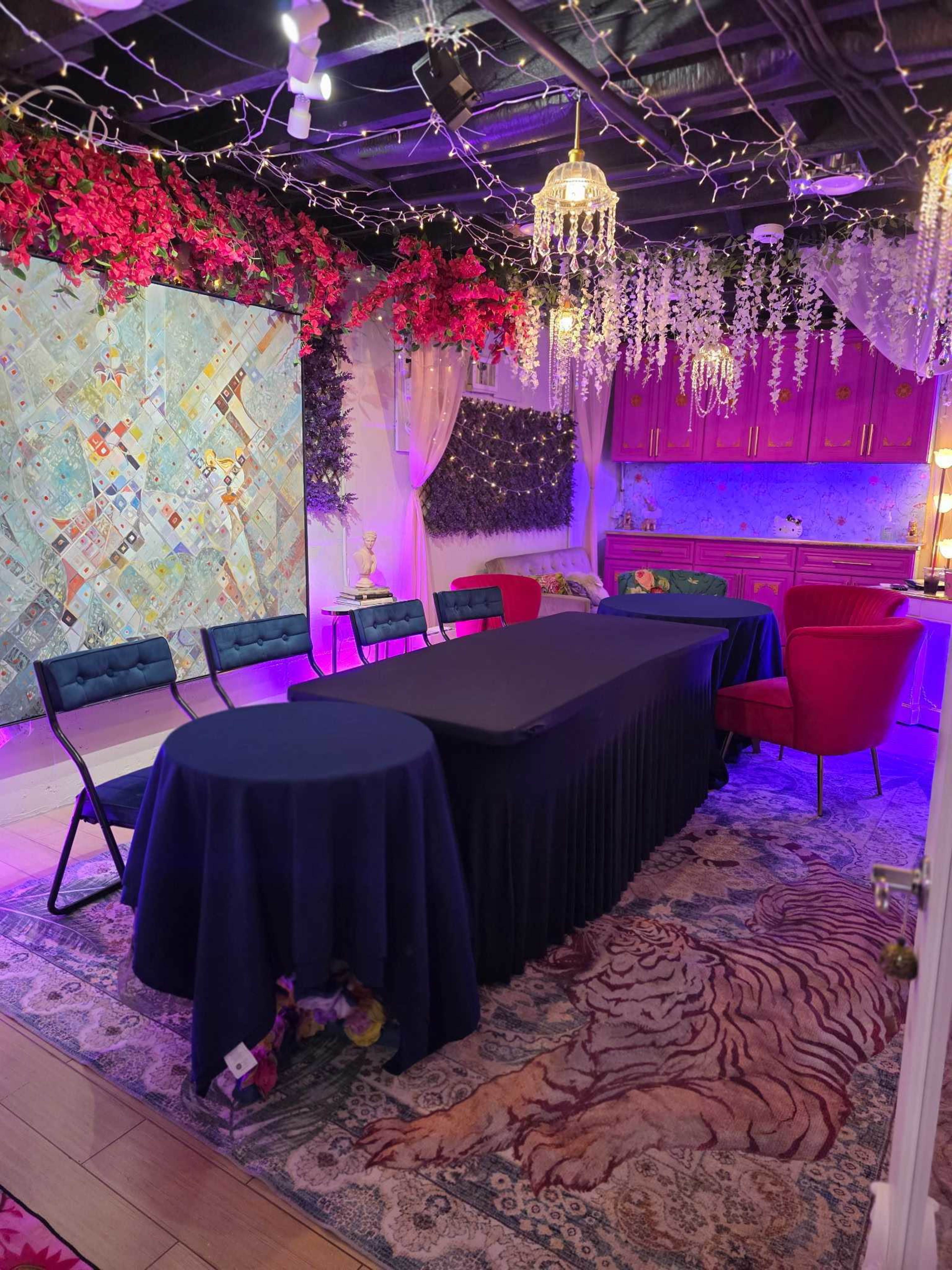 The image shows a decorated indoor space featuring a large table covered with a black tablecloth, surrounded by chairs, with colorful walls and ambient lighting.