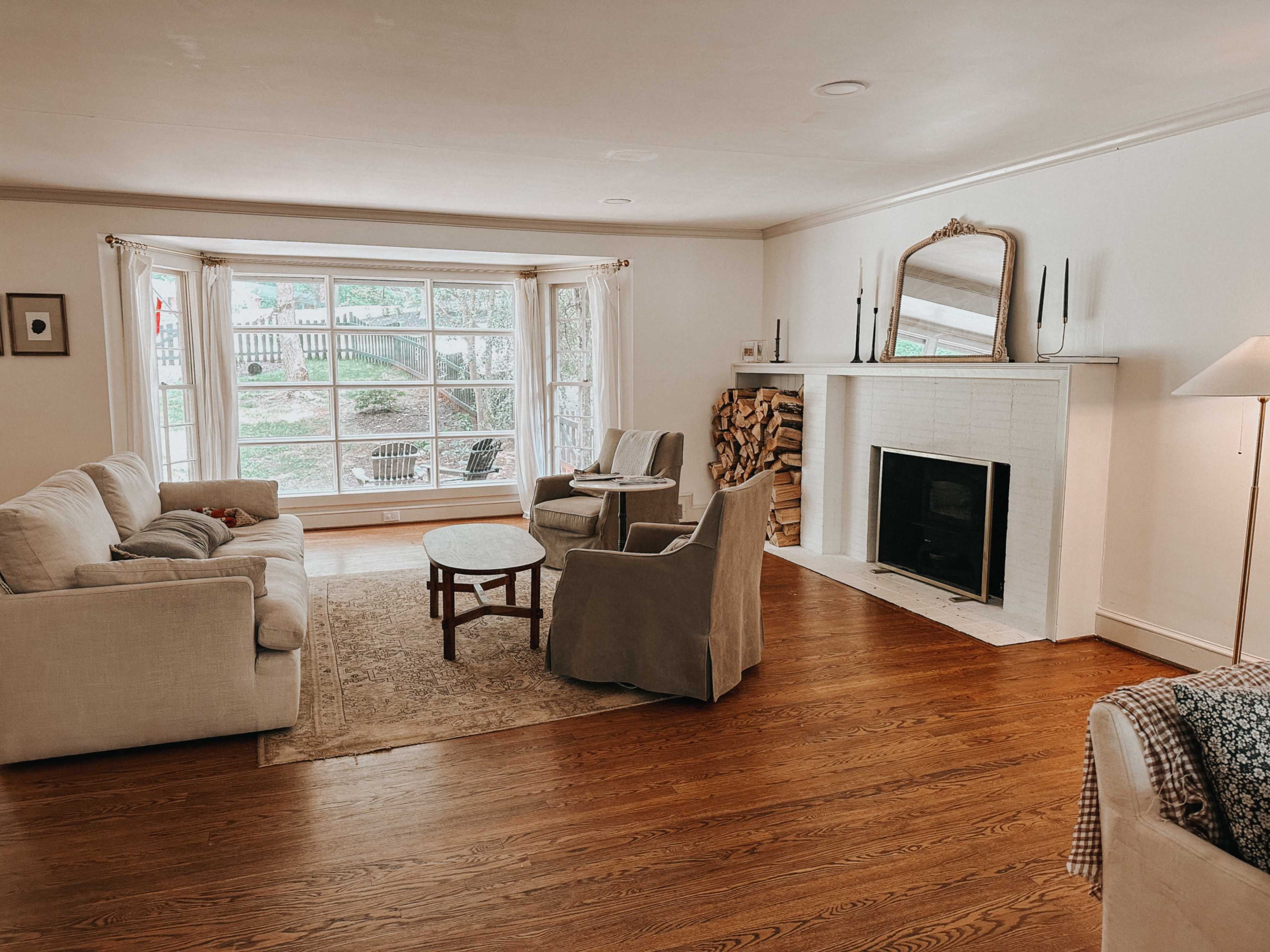 A cozy living room with a fireplace, large windows, a light-colored sofa, two armchairs, a small round table, and a stack of firewood beside the fireplace.