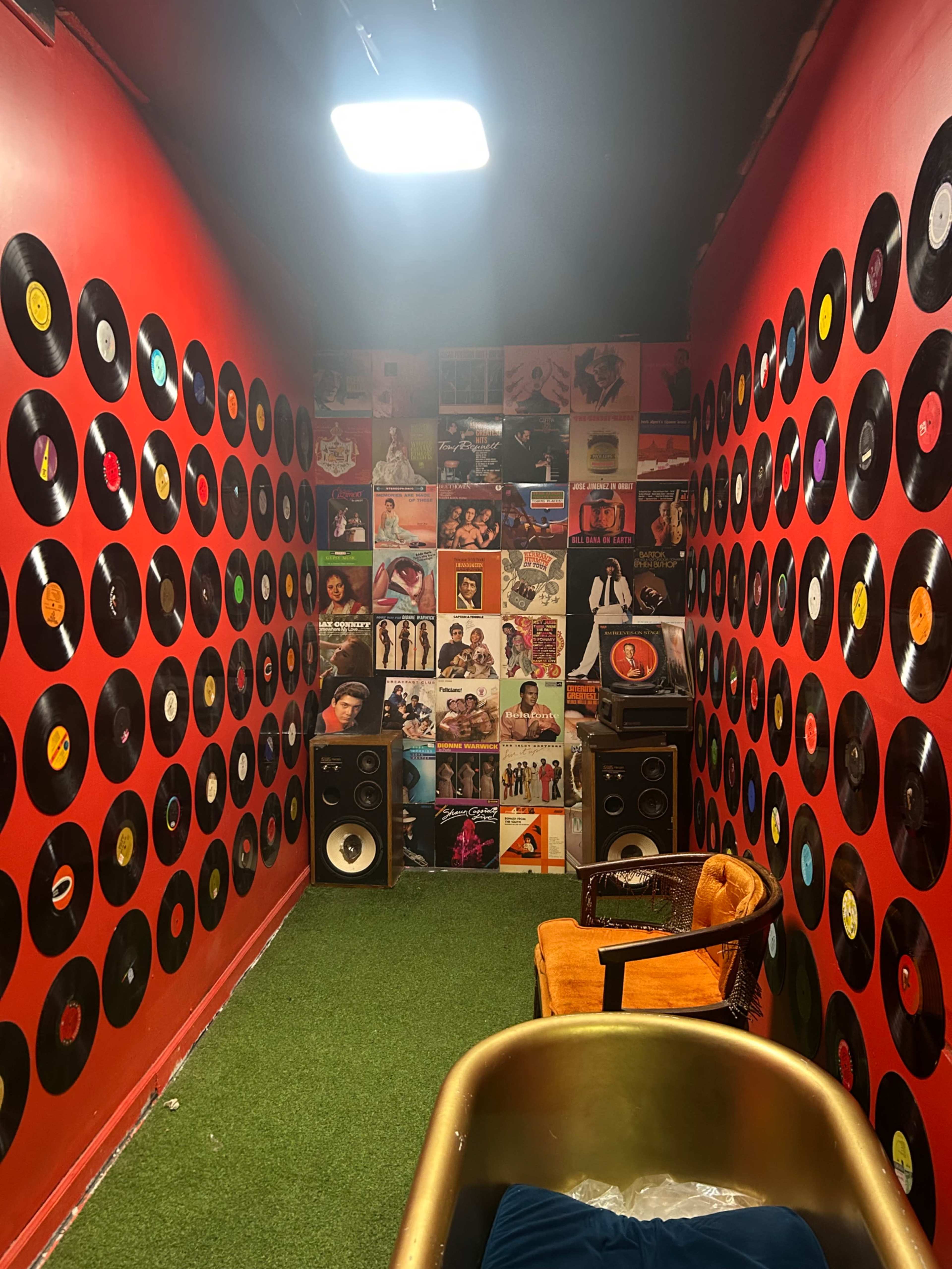The image shows a small, vibrant room with red walls adorned with numerous vinyl records and posters, featuring speakers and a yellow chair on a green carpet.