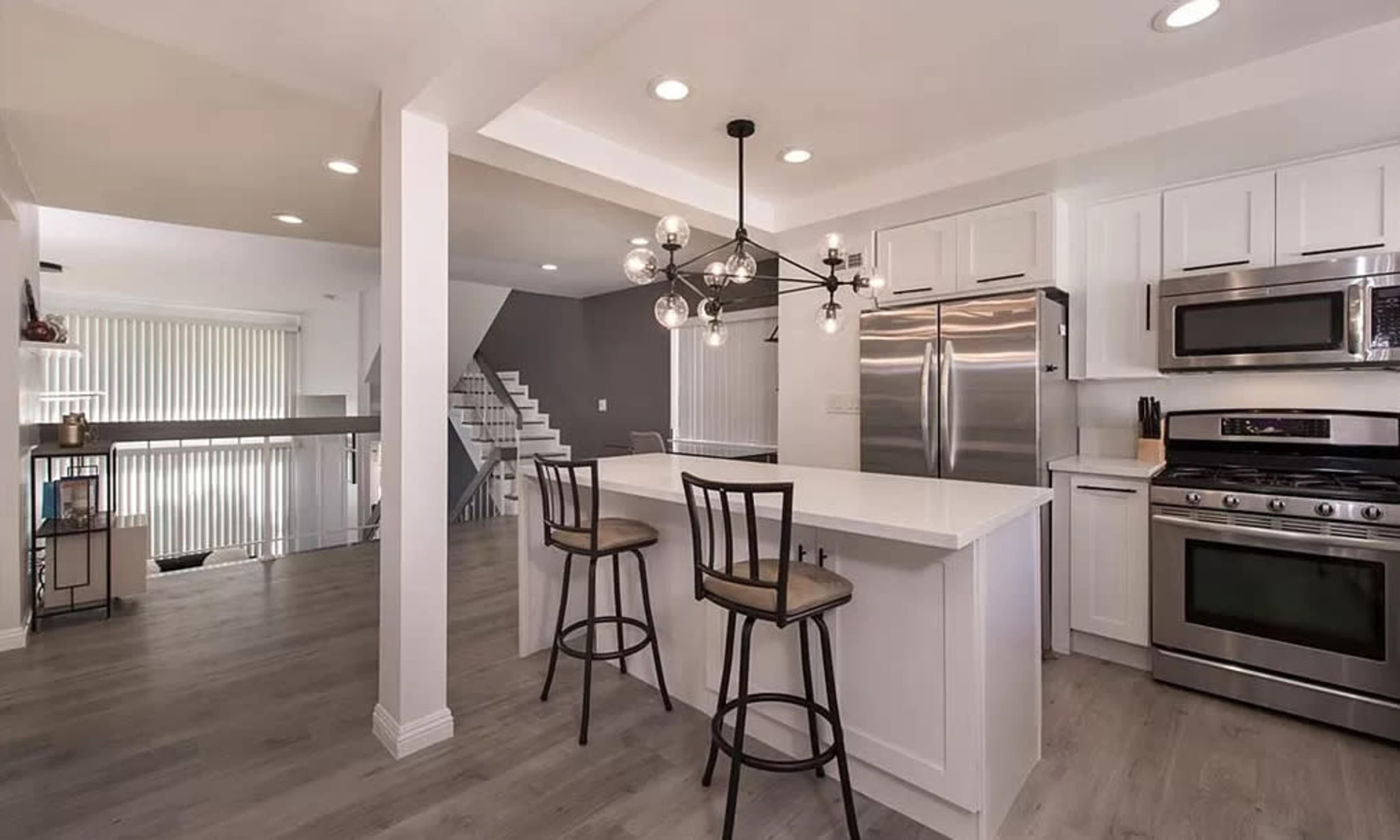 A modern kitchen with a large island, stainless steel appliances, and two bar stools, adjacent to a staircase and an open living area.