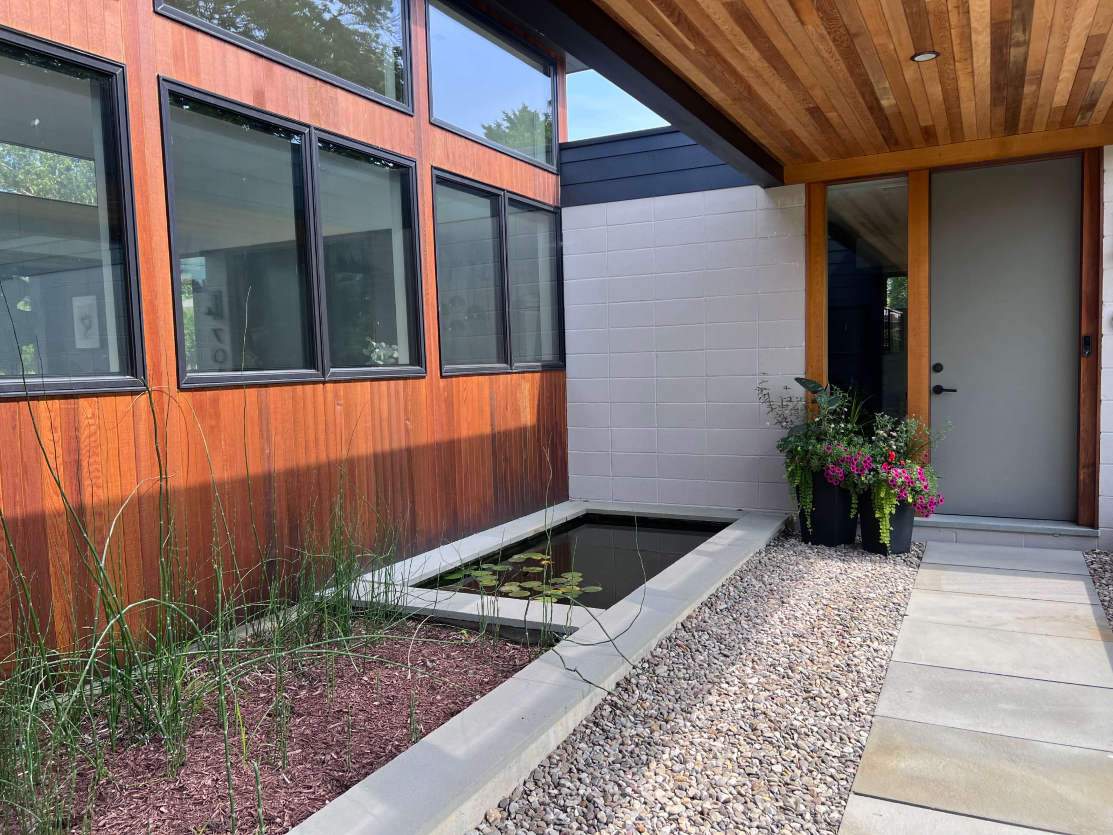 The image shows a modern entrance with a pond, landscaped plants, and a wooden overhang leading to a front door.