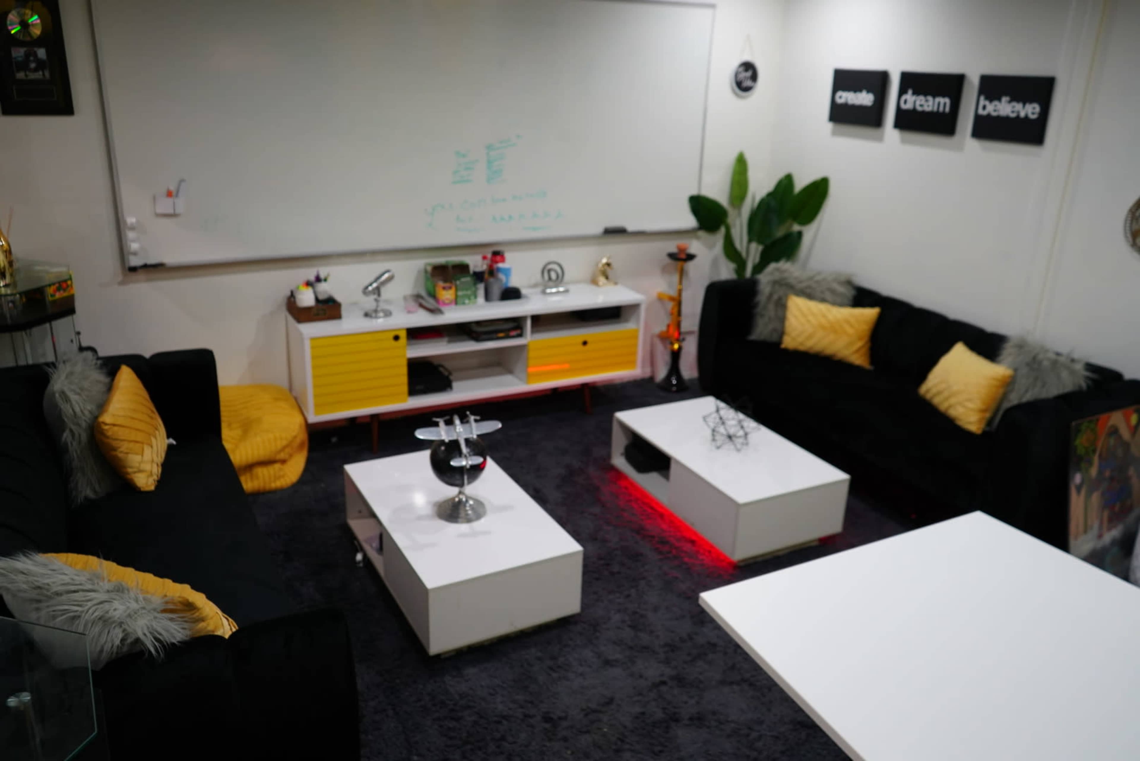 A modern lounge area with black sofas, white coffee tables, and a whiteboard on the wall, adorned with decorative plants and motivational wall art.