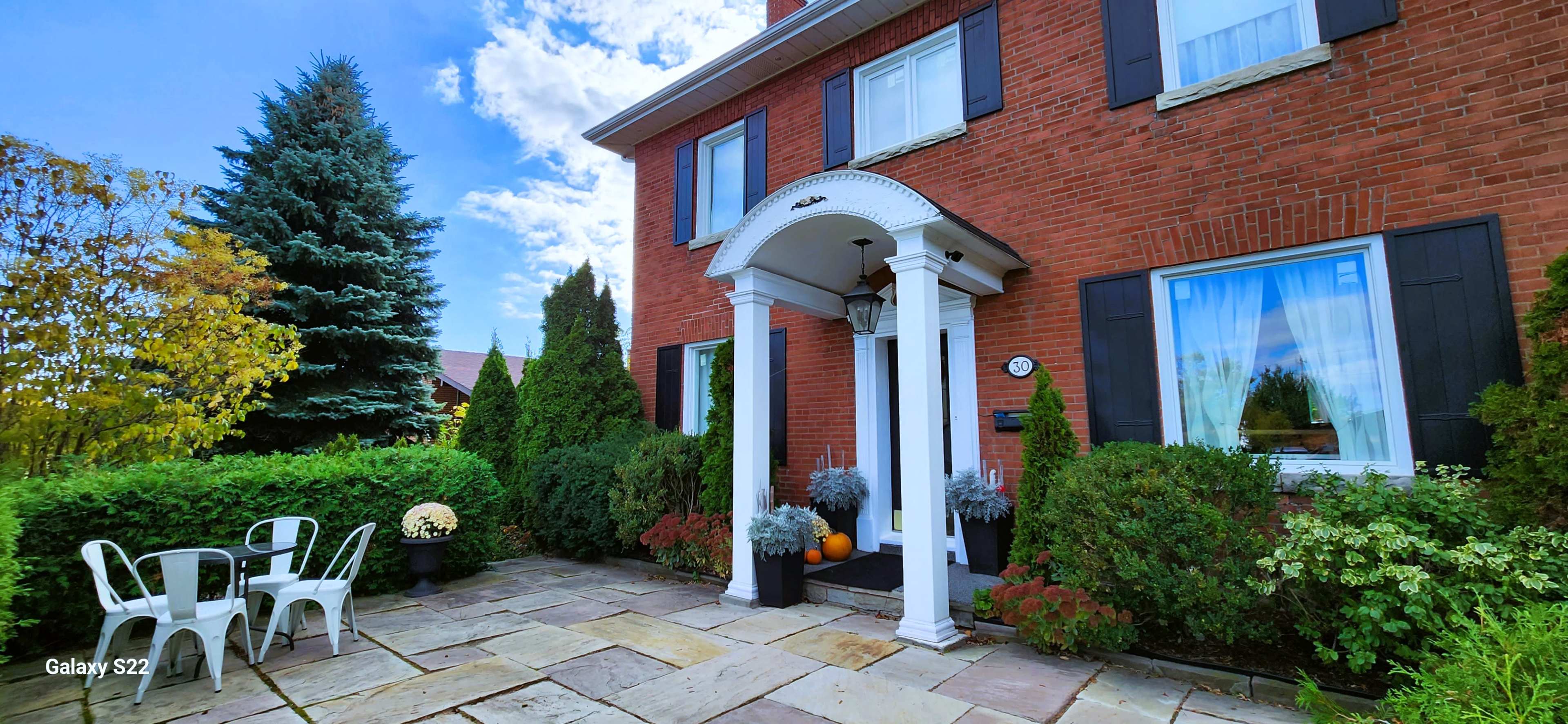 A brick house with white columns at the entrance, surrounded by greenery and a stone patio with two white chairs and potted plants.