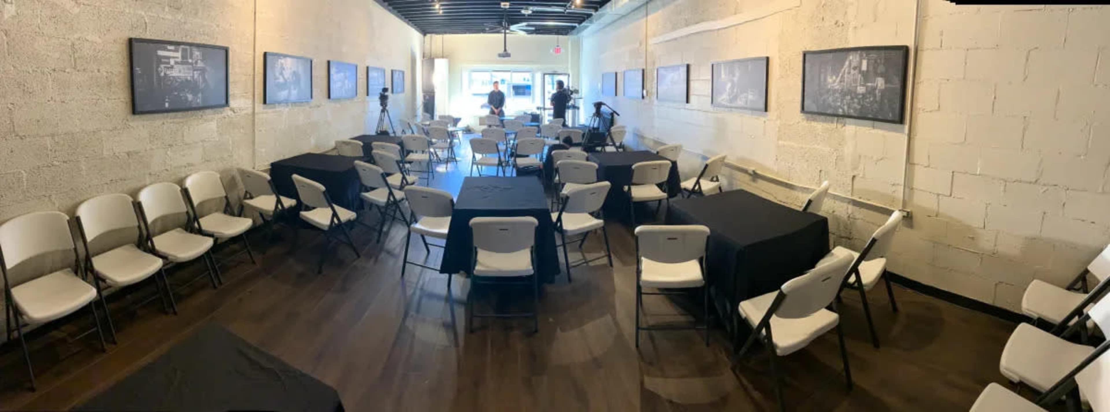 A spacious interior room arranged with rows of white chairs and black tables, with black-and-white photographs hanging on the walls.
