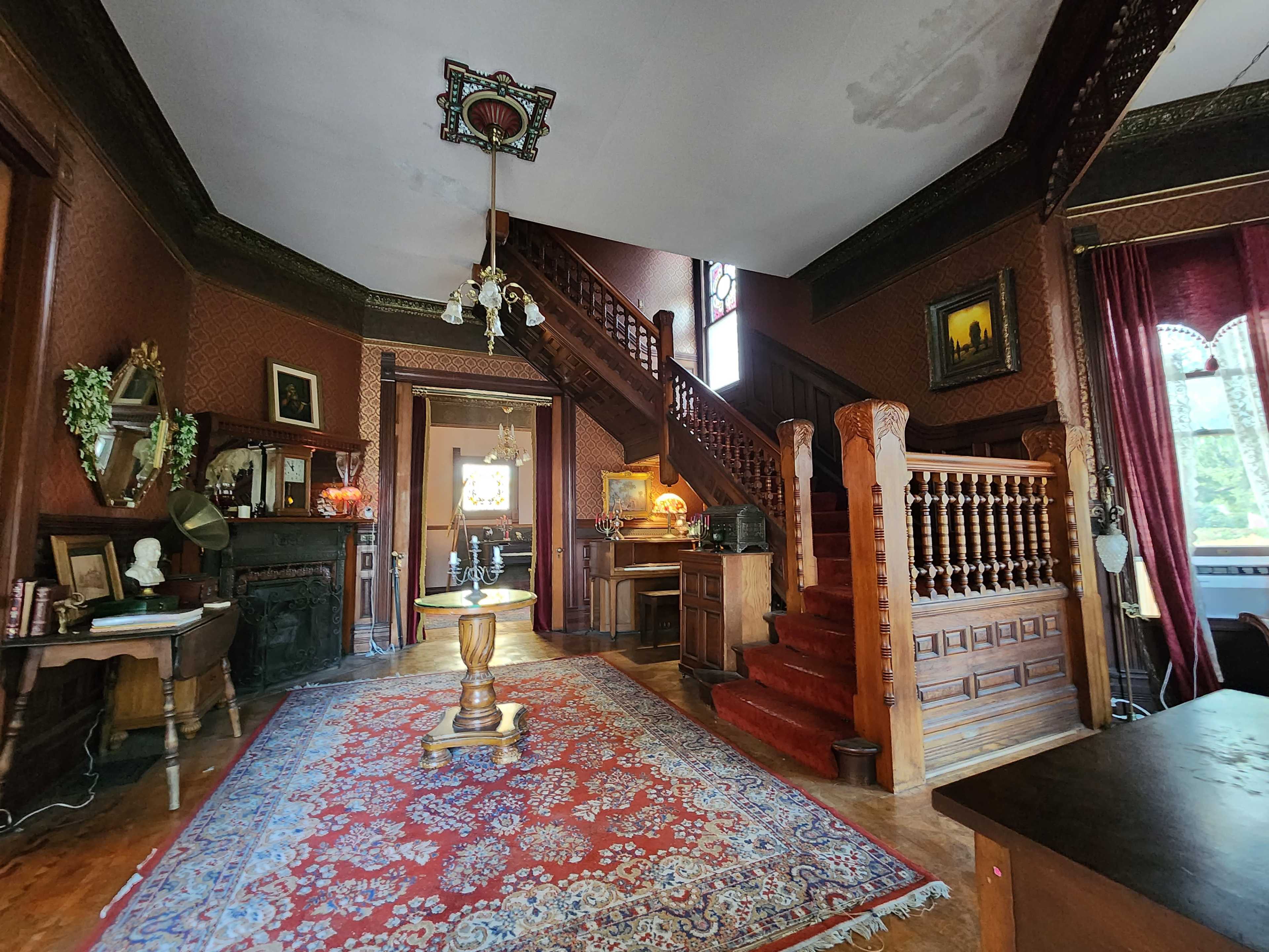 A vintage interior of a home featuring a staircase, ornate woodwork, a large patterned rug, and period furnishings.