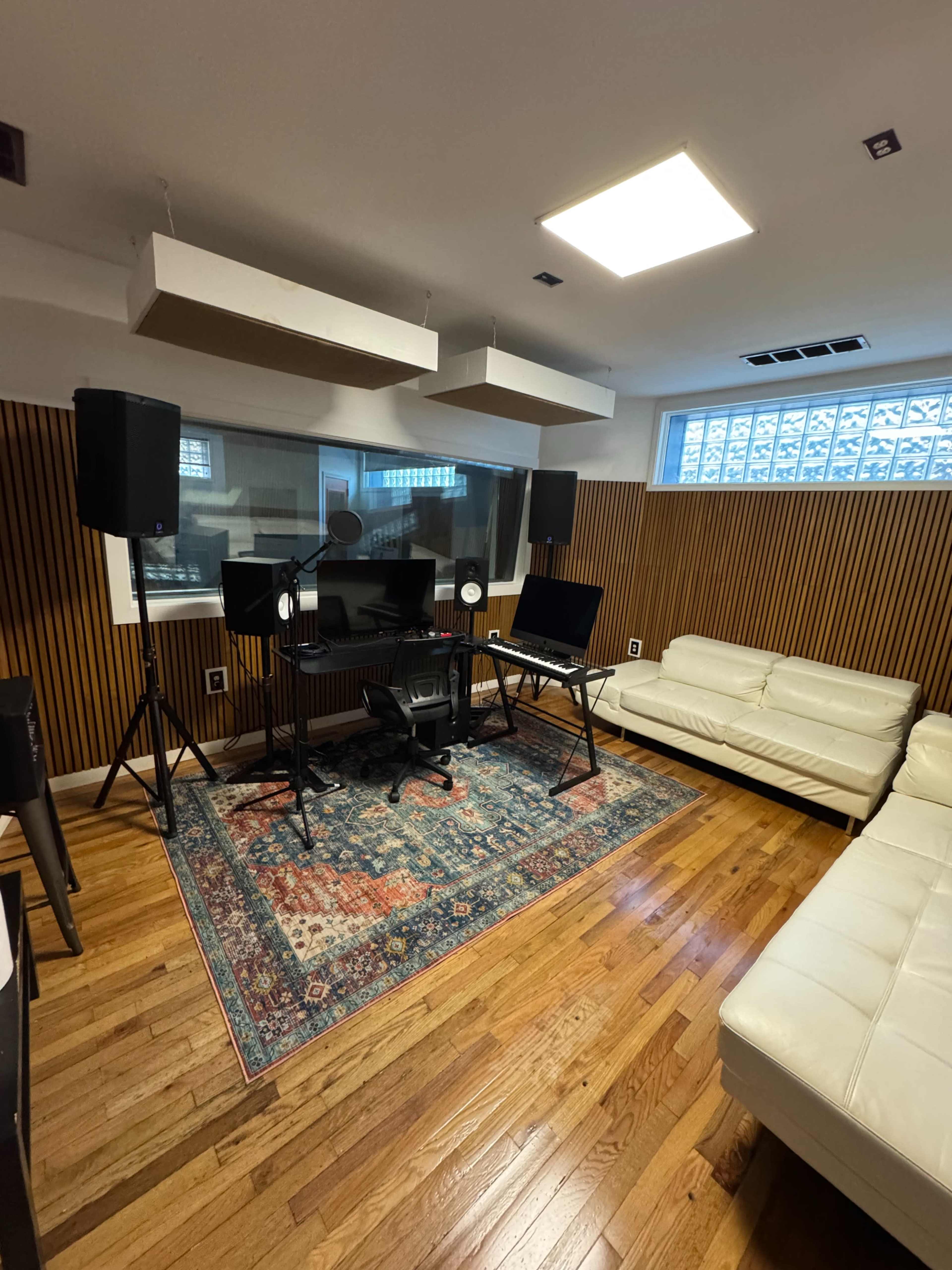 The image depicts a music studio featuring a mixing desk, two monitor speakers, a keyboard stand, and a white sofa, all set against wood-paneled walls.