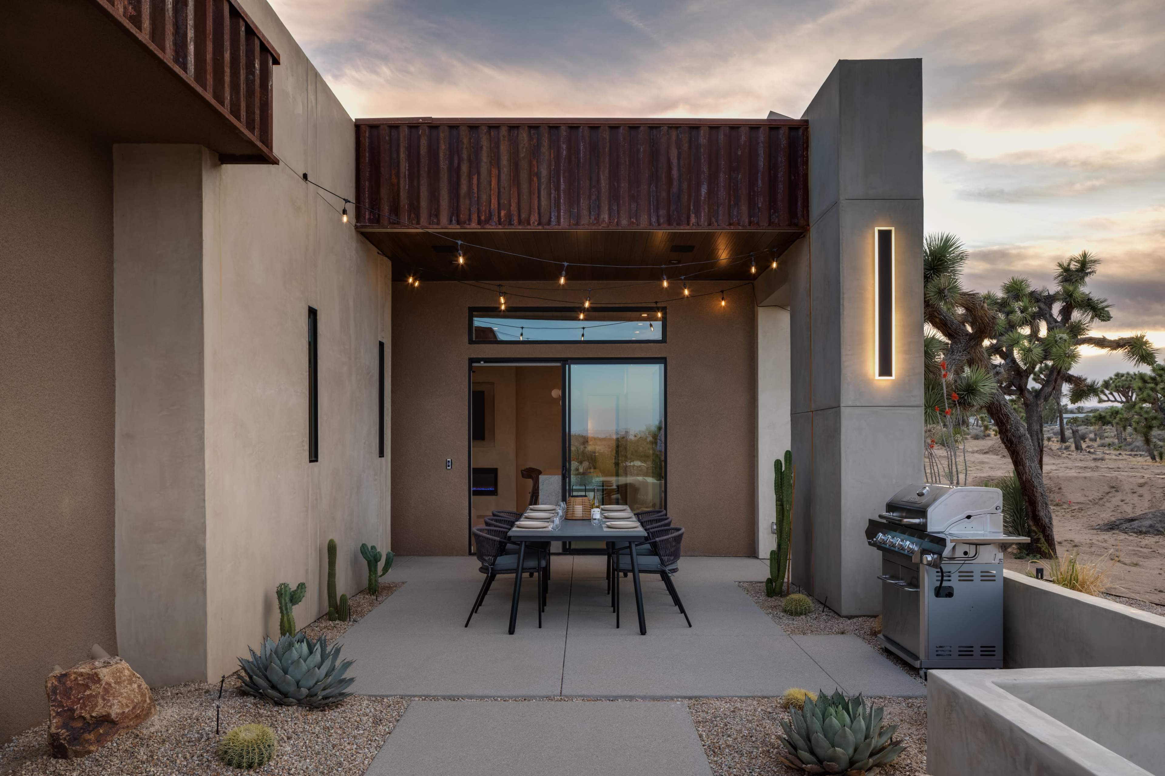 A modern outdoor dining area with a table set for meals is situated in front of a contemporary desert home, surrounded by cacti and succulents.