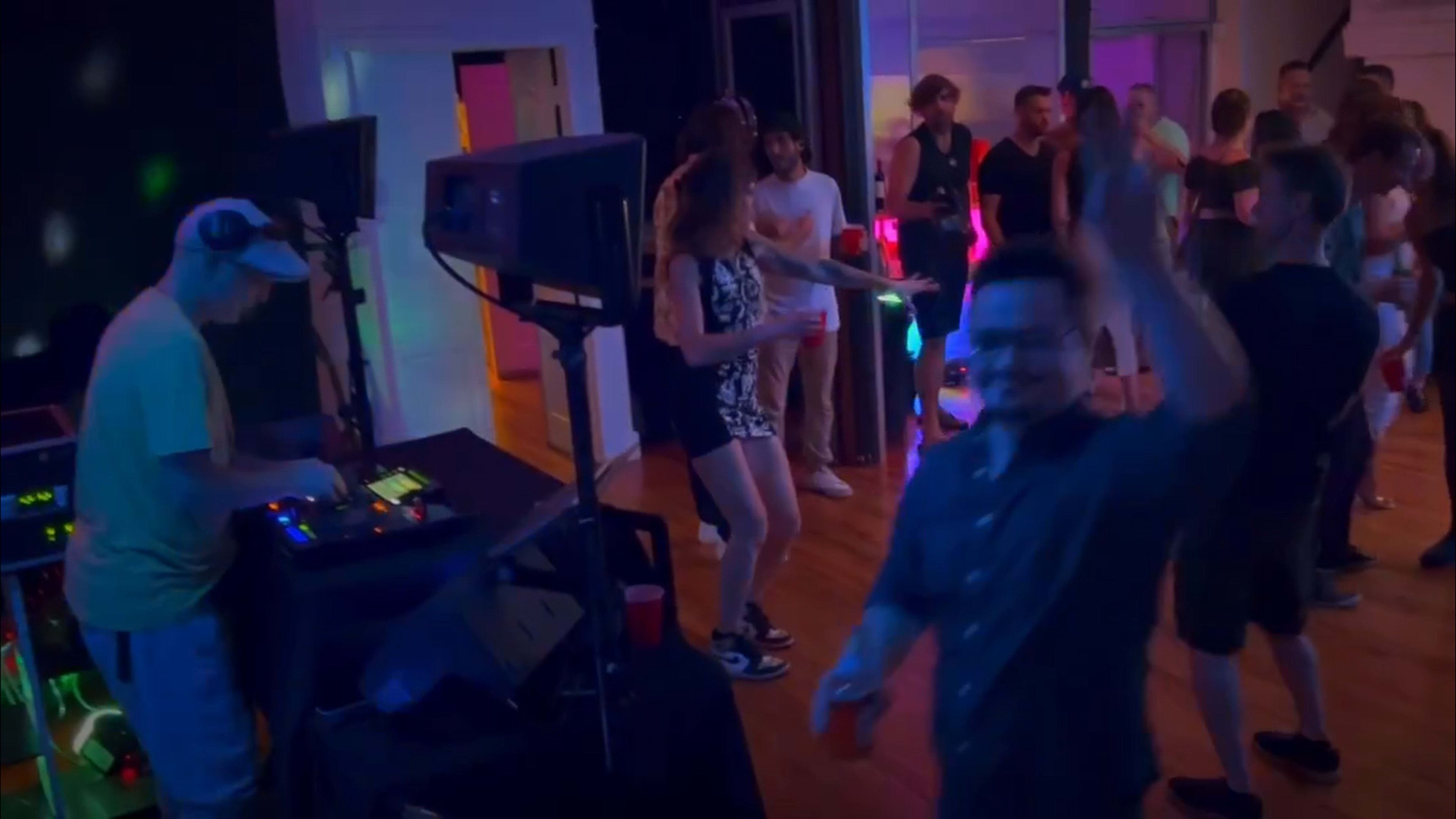 A DJ performs in front of an energized crowd dancing under colorful lights in a lively indoor venue.