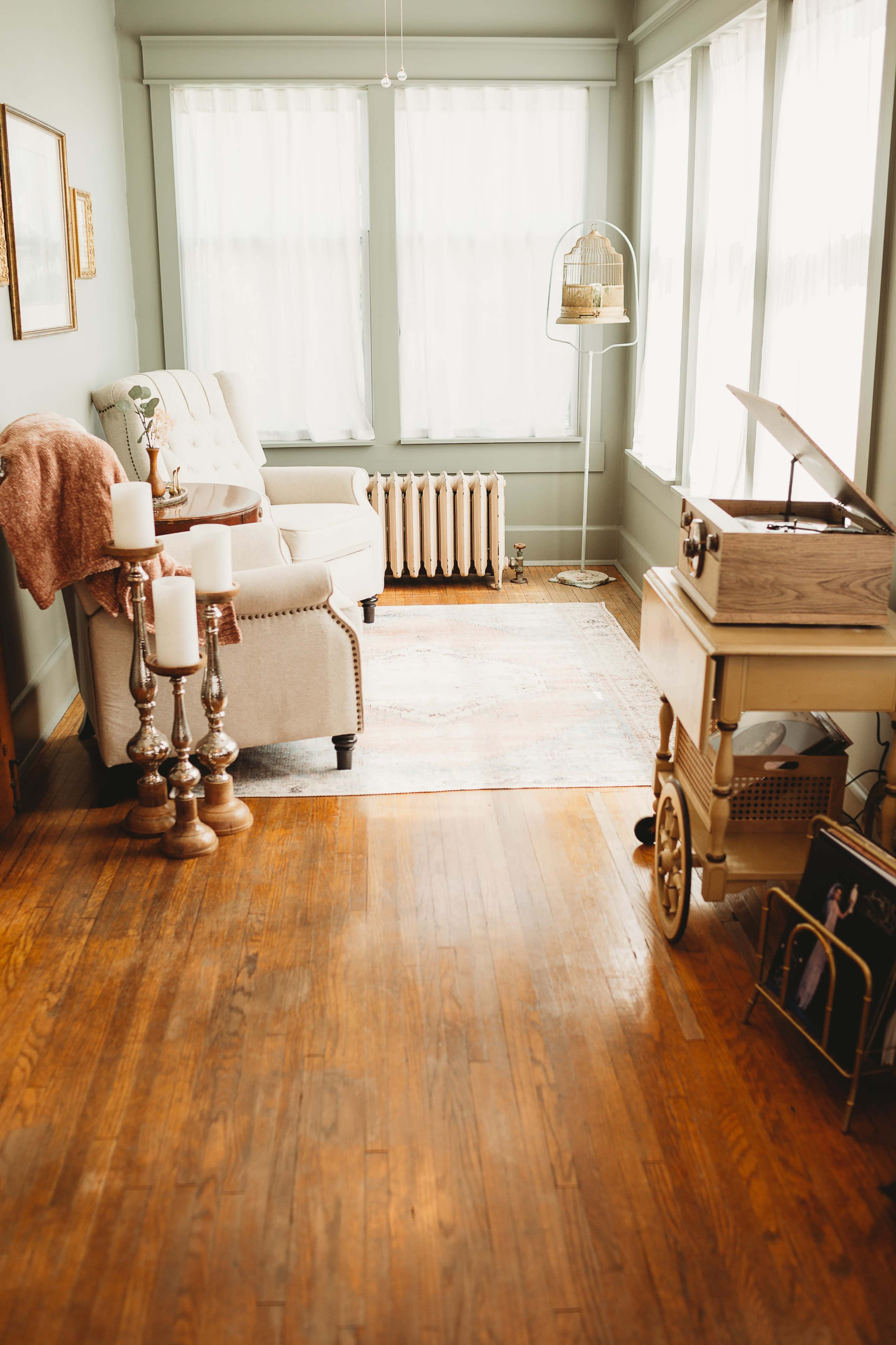 A well-lit living room features a comfortable armchair, a wooden record player on a cart, and a series of elegant candle holders beside a radiator.