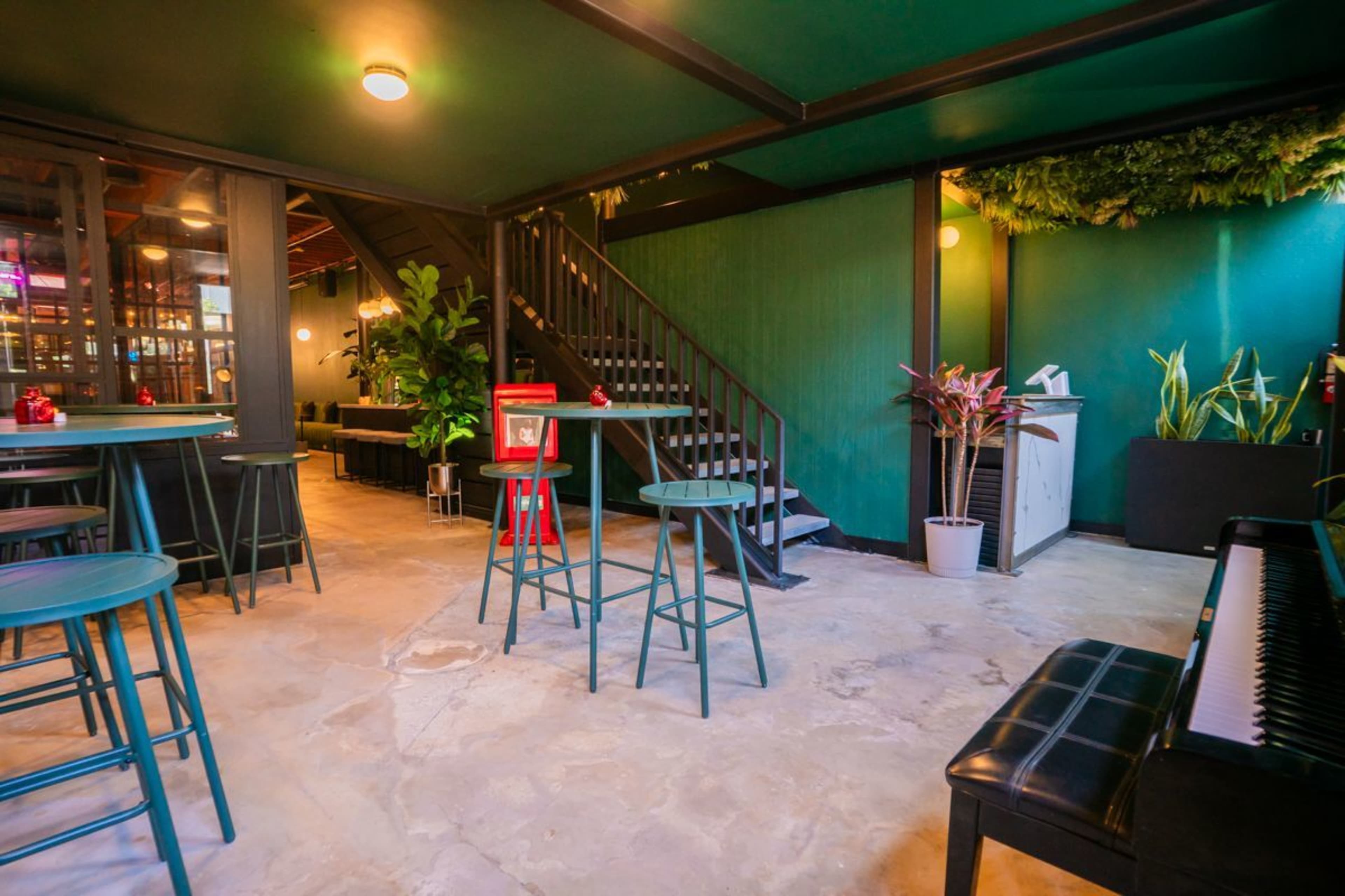 The image shows a contemporary indoor space with green walls, dark furniture, and a staircase, featuring plants and a piano in the corner.