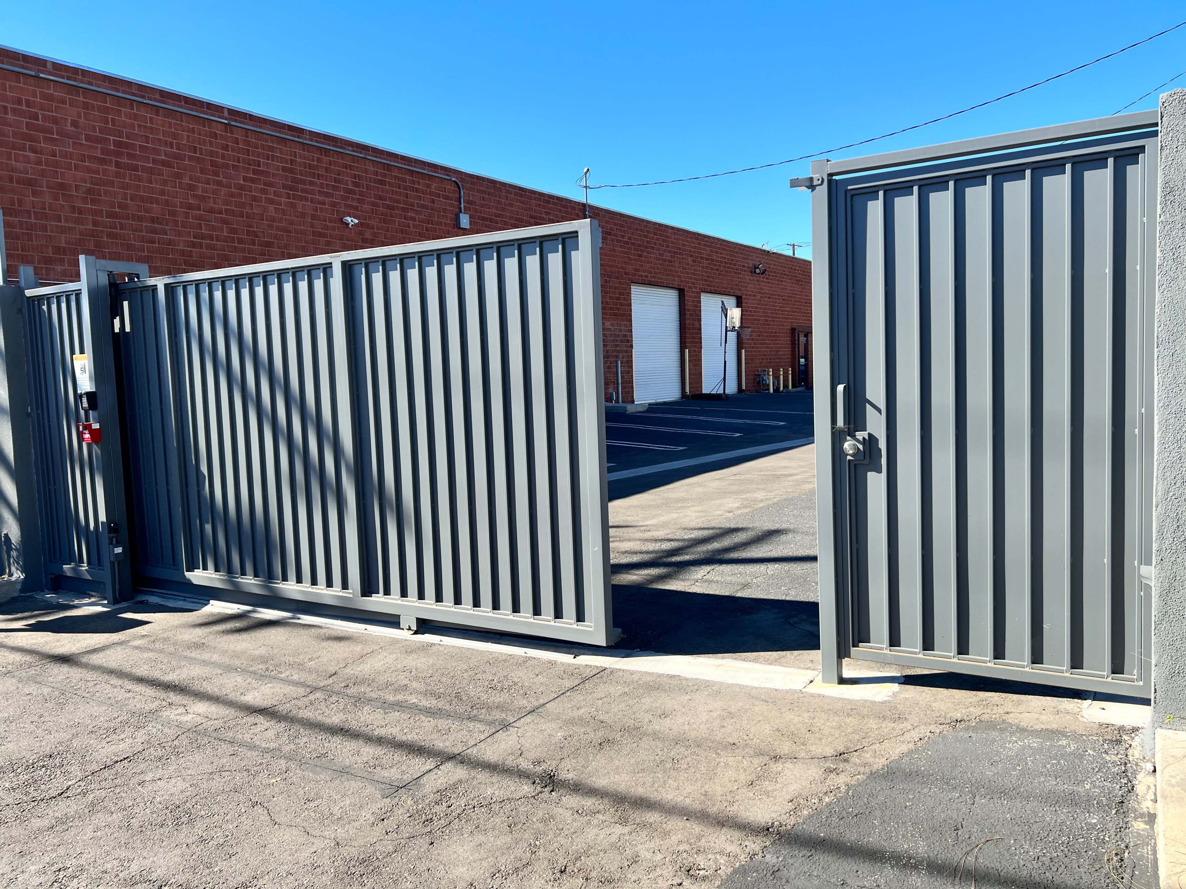 A pair of gray metal gates stands open, leading to a paved parking area behind a building with a brick facade.