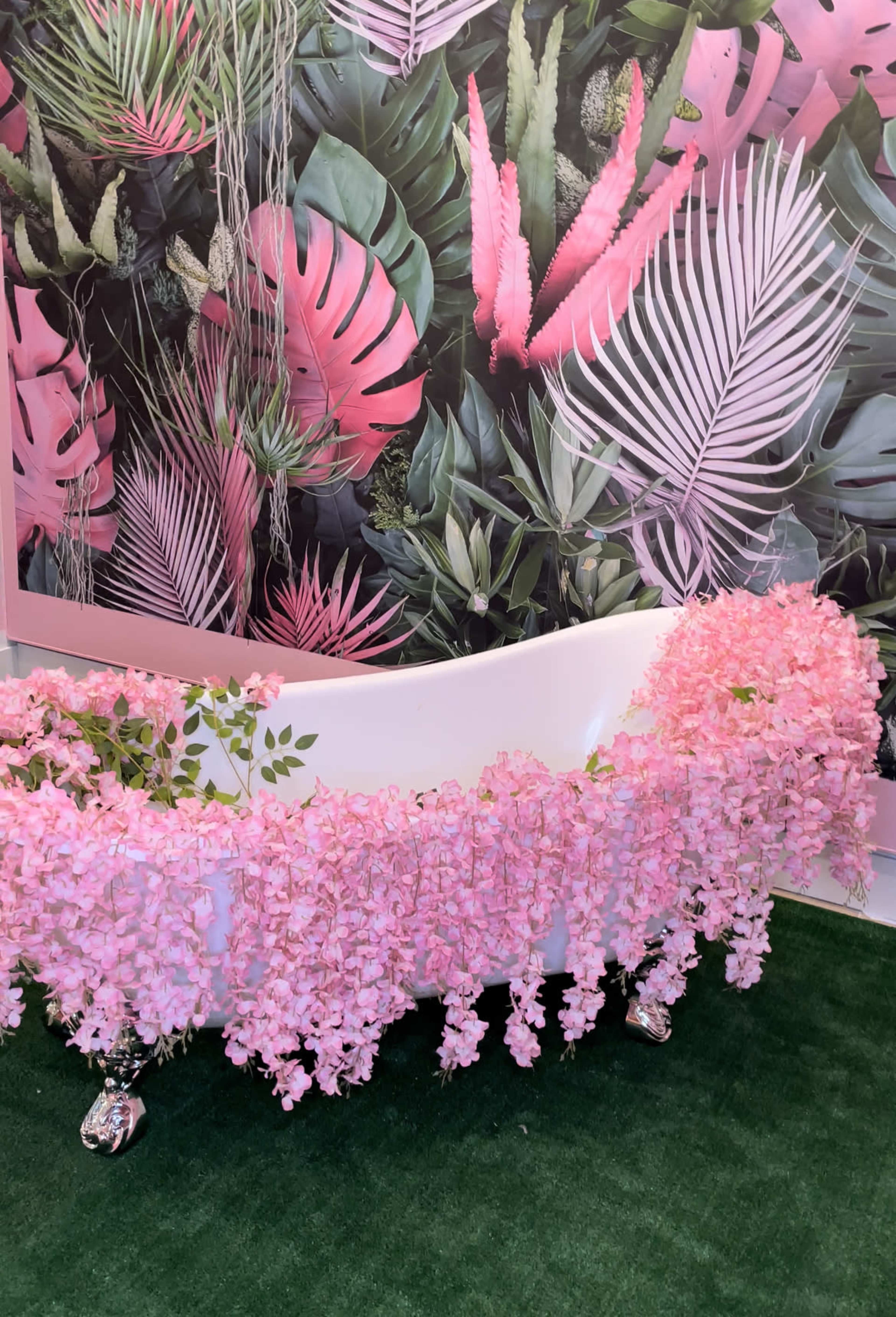A white clawfoot bathtub is adorned with cascading pink flowers against a backdrop of tropical foliage and pink leaves.