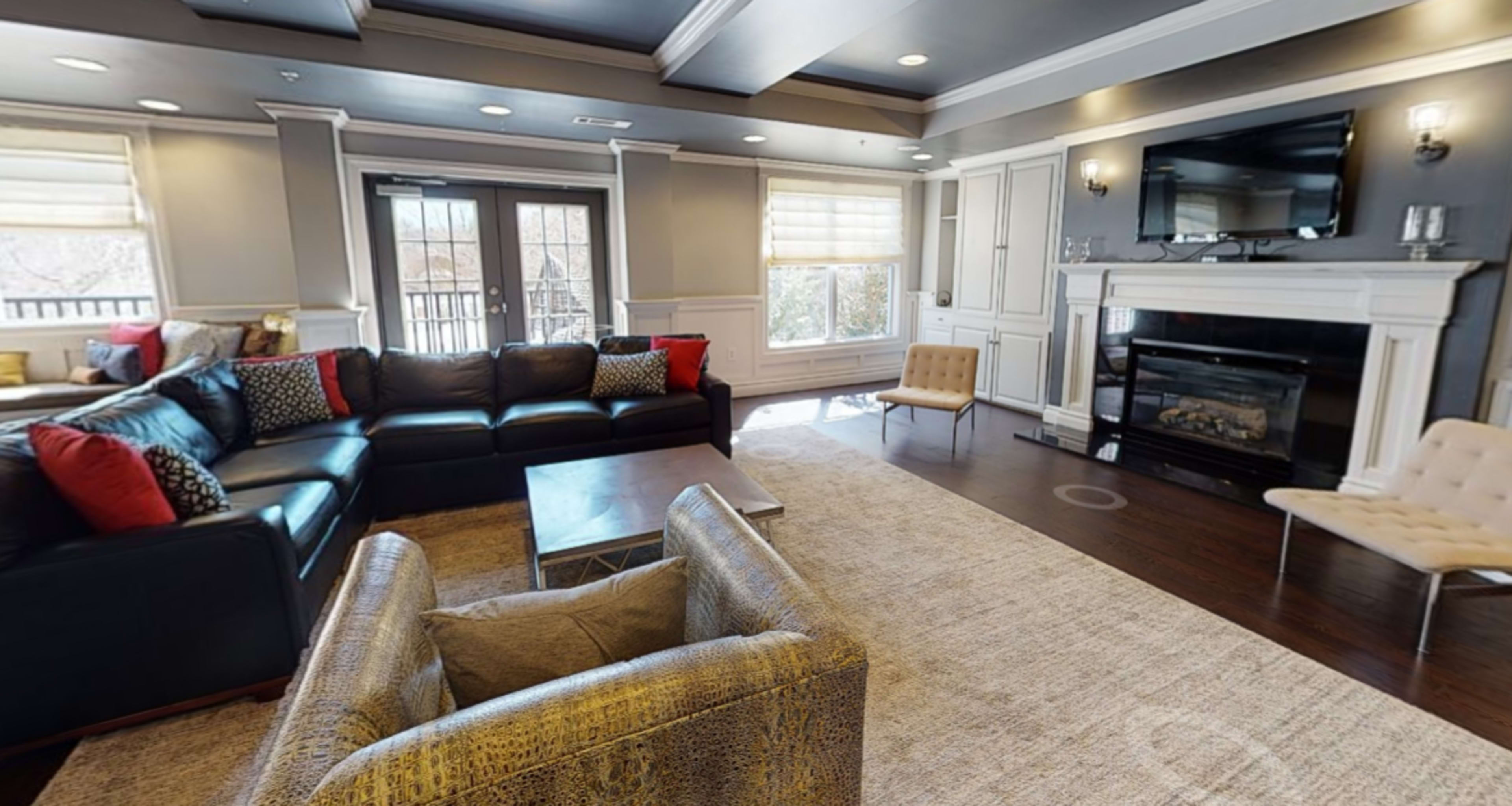 The image shows a modern living room with a black sectional sofa, a coffee table, and a fireplace, all surrounded by neutral-toned walls and large windows letting in natural light.