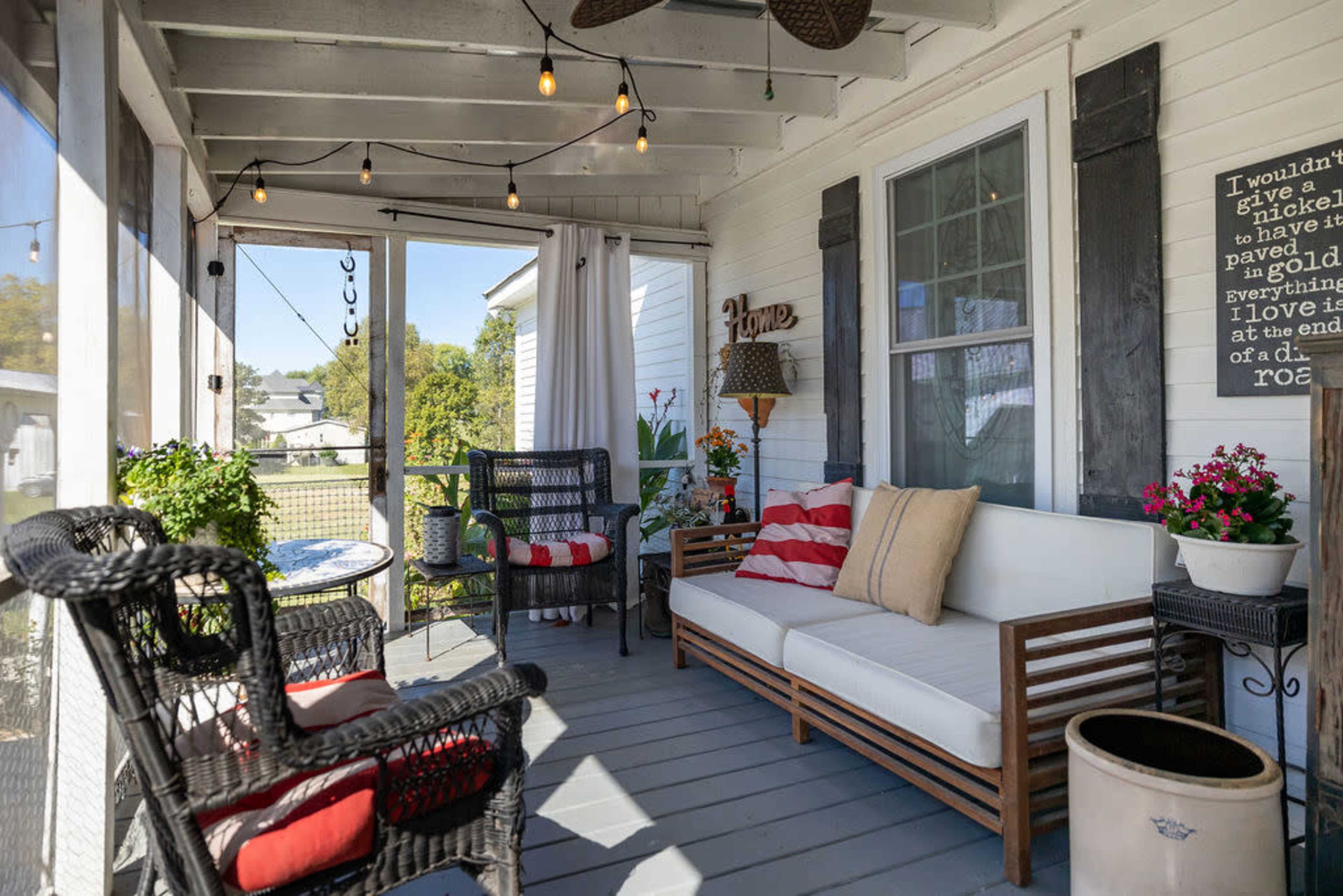 The image shows a screened porch with a white sofa, black wicker chairs, potted plants, and decorative string lights.