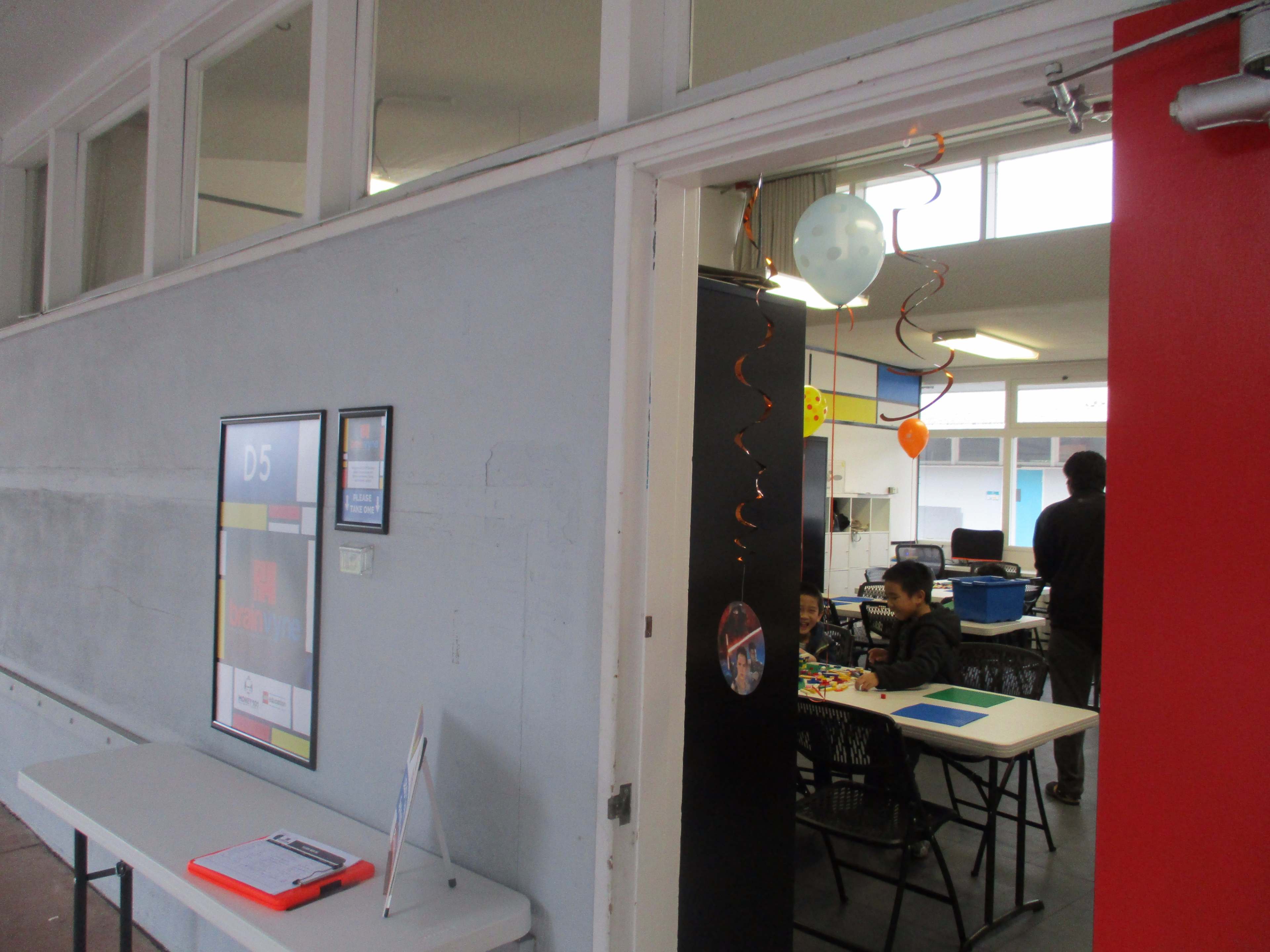 A table is set up in a hallway with a framed poster, while a classroom is visible in the background with a student sitting at a table.