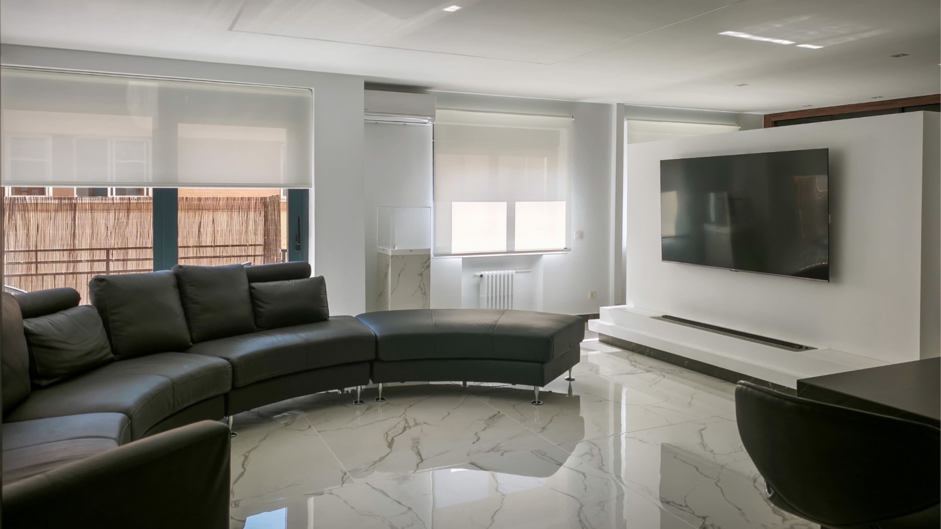 The image shows a modern living room featuring a curved black leather sectional sofa, a large flat-screen TV mounted on a white wall, and glossy marble flooring.