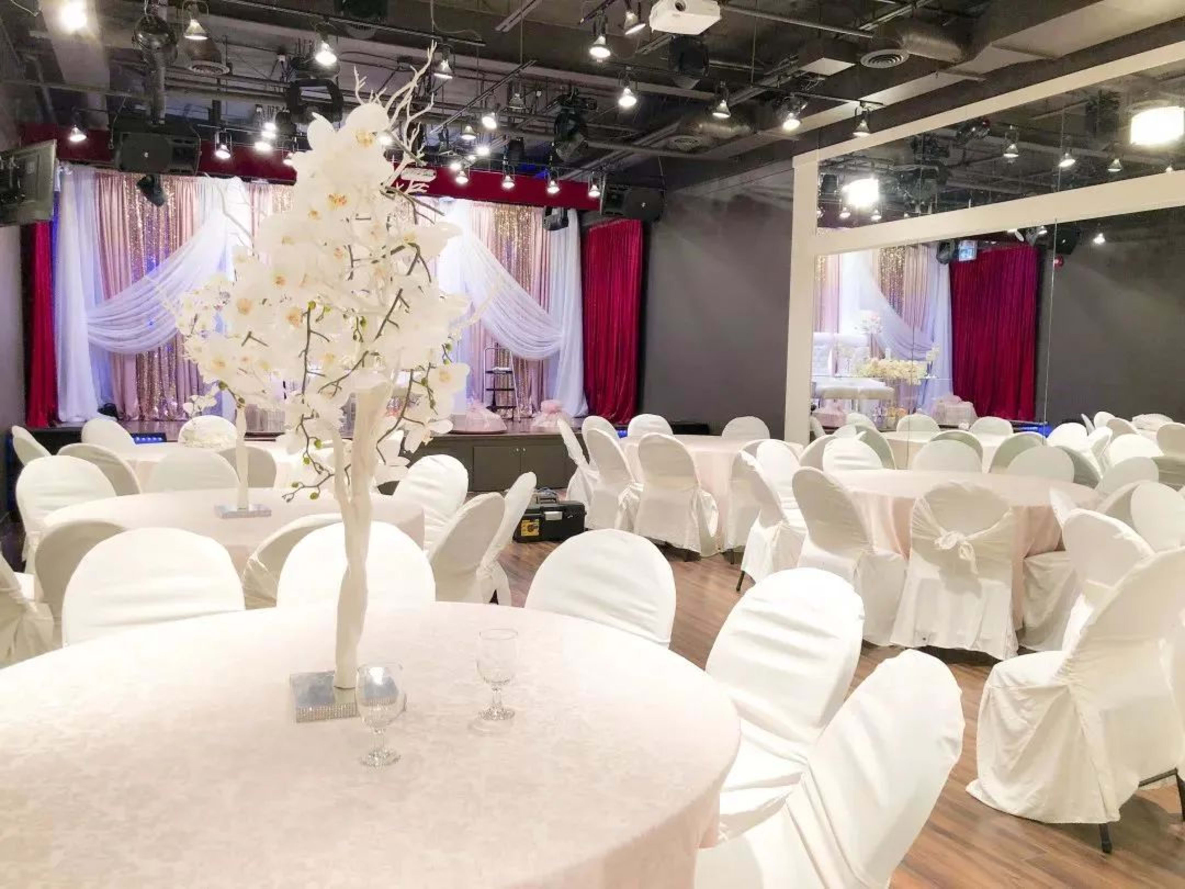 A banquet hall set up with round tables covered in white tablecloths and adorned with white floral centerpieces, accompanied by a stage in the background.