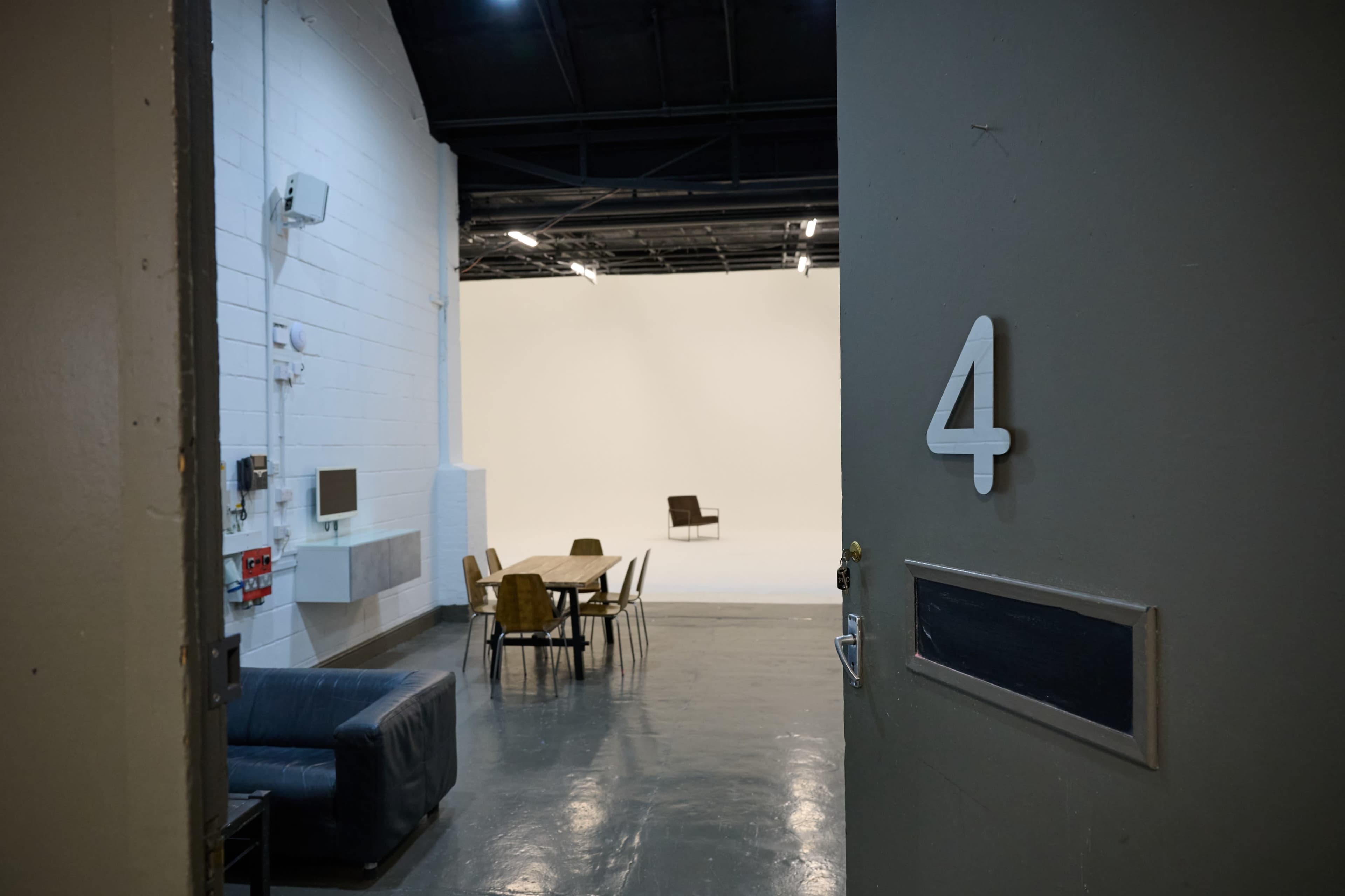 The image shows an open door with the number 4, revealing a spacious studio area with a white backdrop and a few chairs arranged in the background.