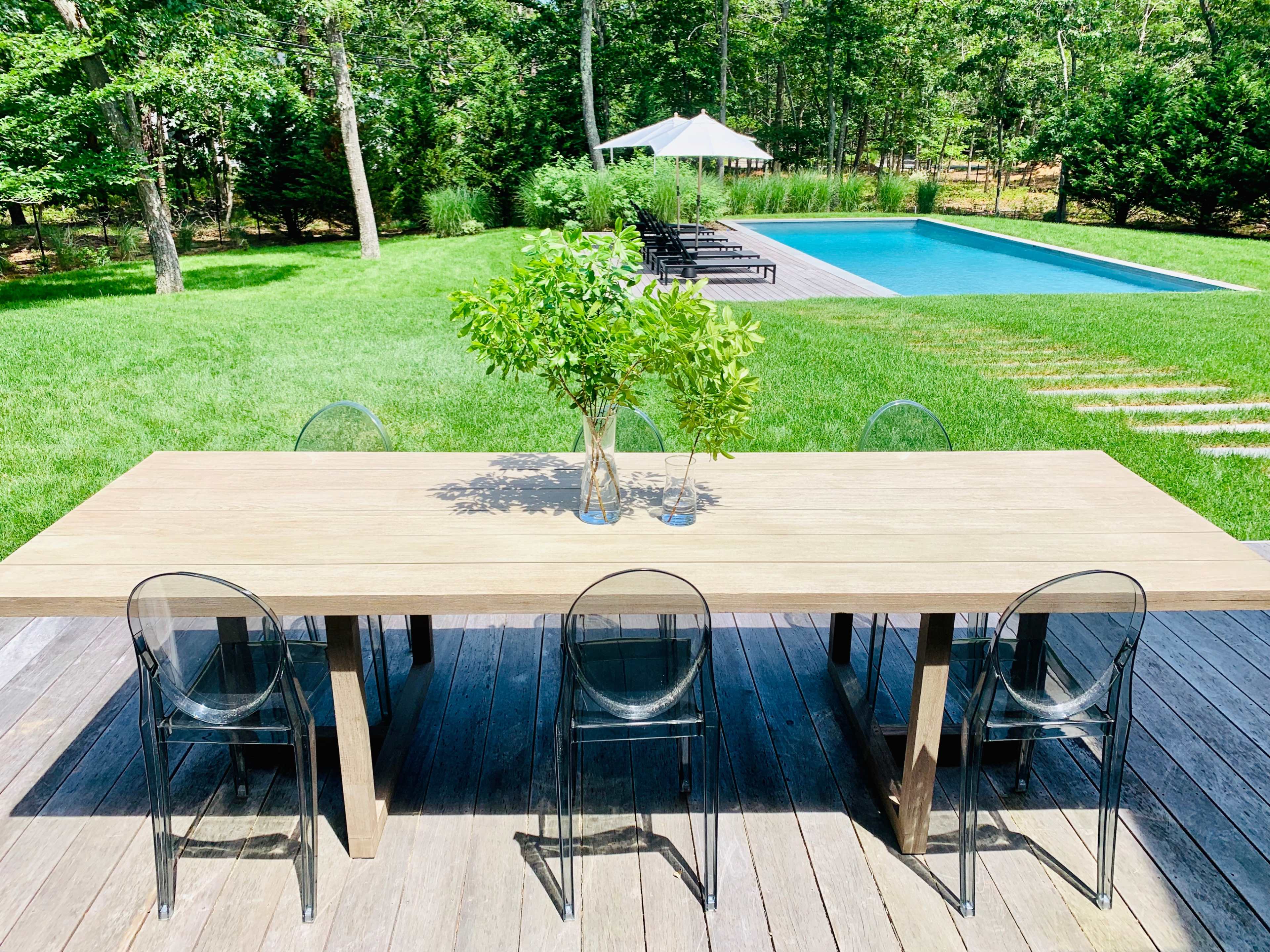 A wooden dining table with transparent chairs is set on a deck, overlooking a lawn with a swimming pool and lounge chairs in the background.