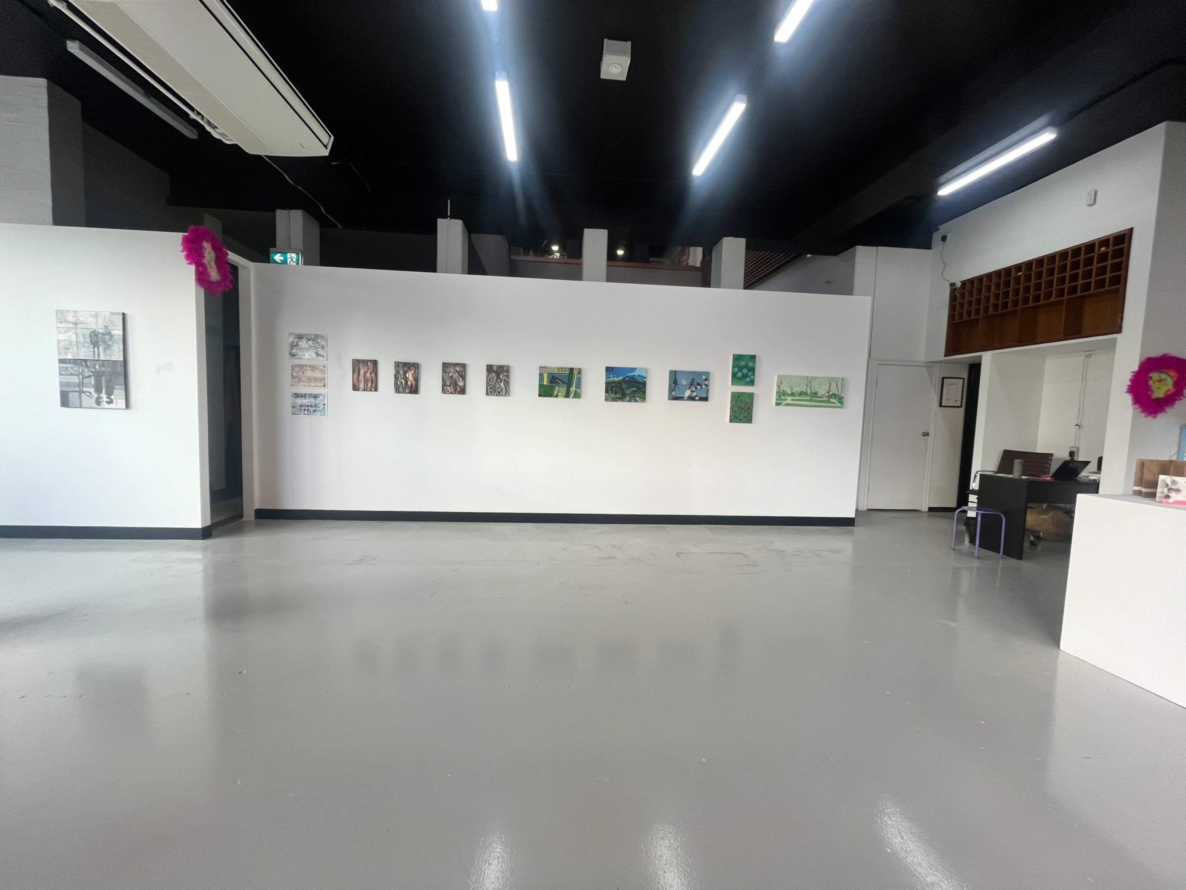 The image shows an art gallery with a white wall displaying several framed artworks, while the floor is polished and the room is brightly lit.
