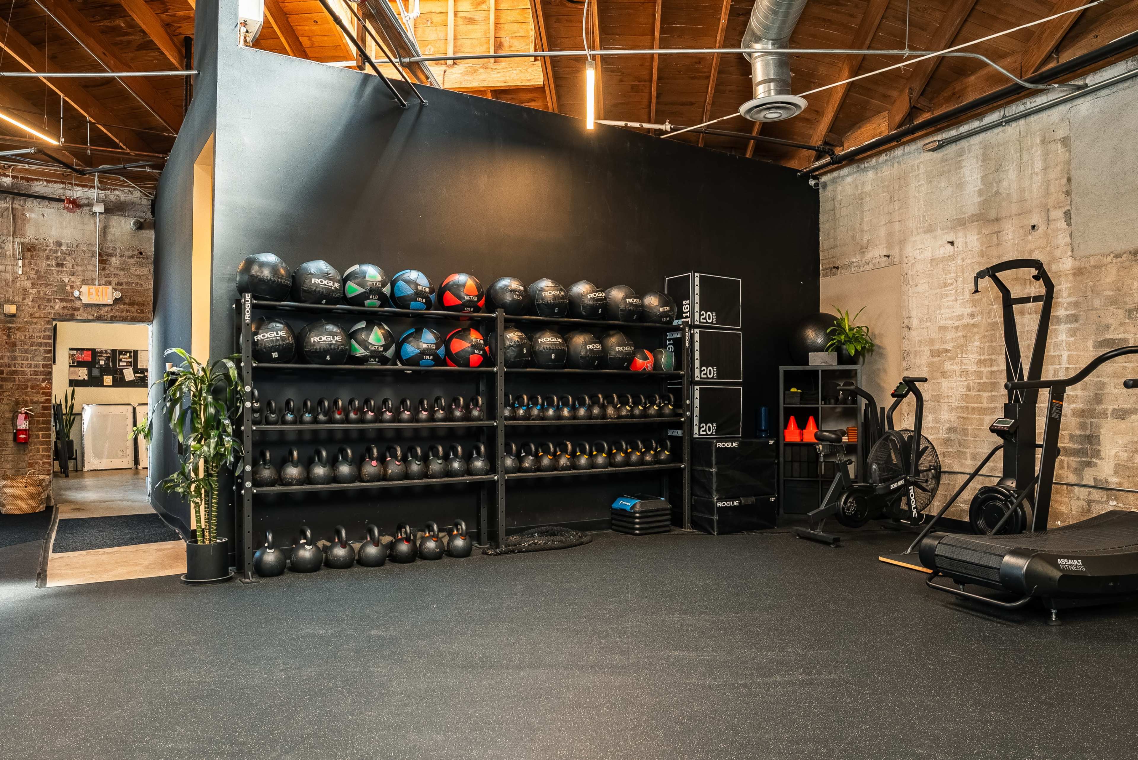The image shows a fitness space with a wall of exercise equipment including kettlebells, medicine balls, and a cardio machine, all set against a brick wall and wooden ceiling.