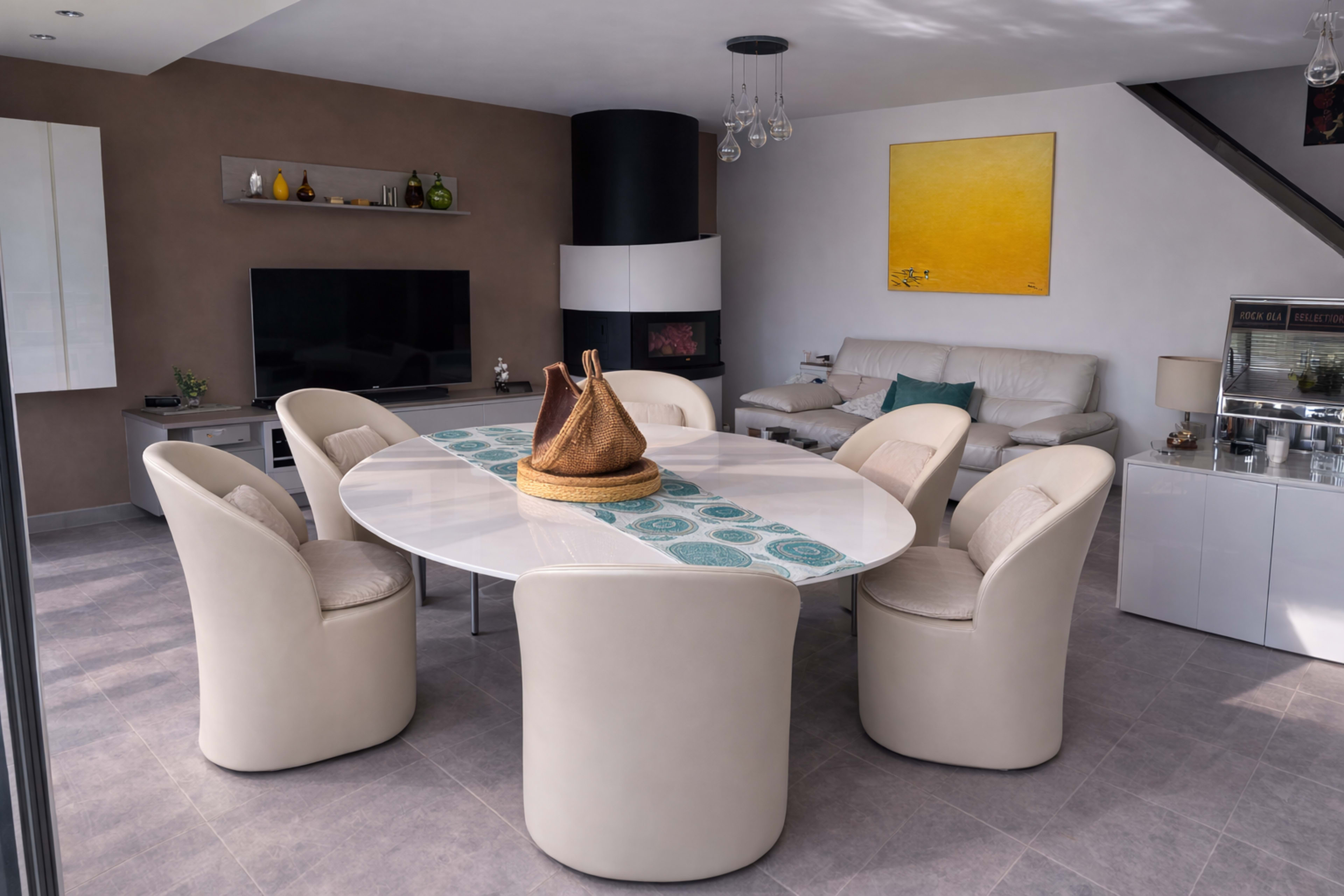 The image shows a modern dining area with a circular white table surrounded by six cream-colored chairs, a television unit, and a beige couch in a stylishly decorated room.