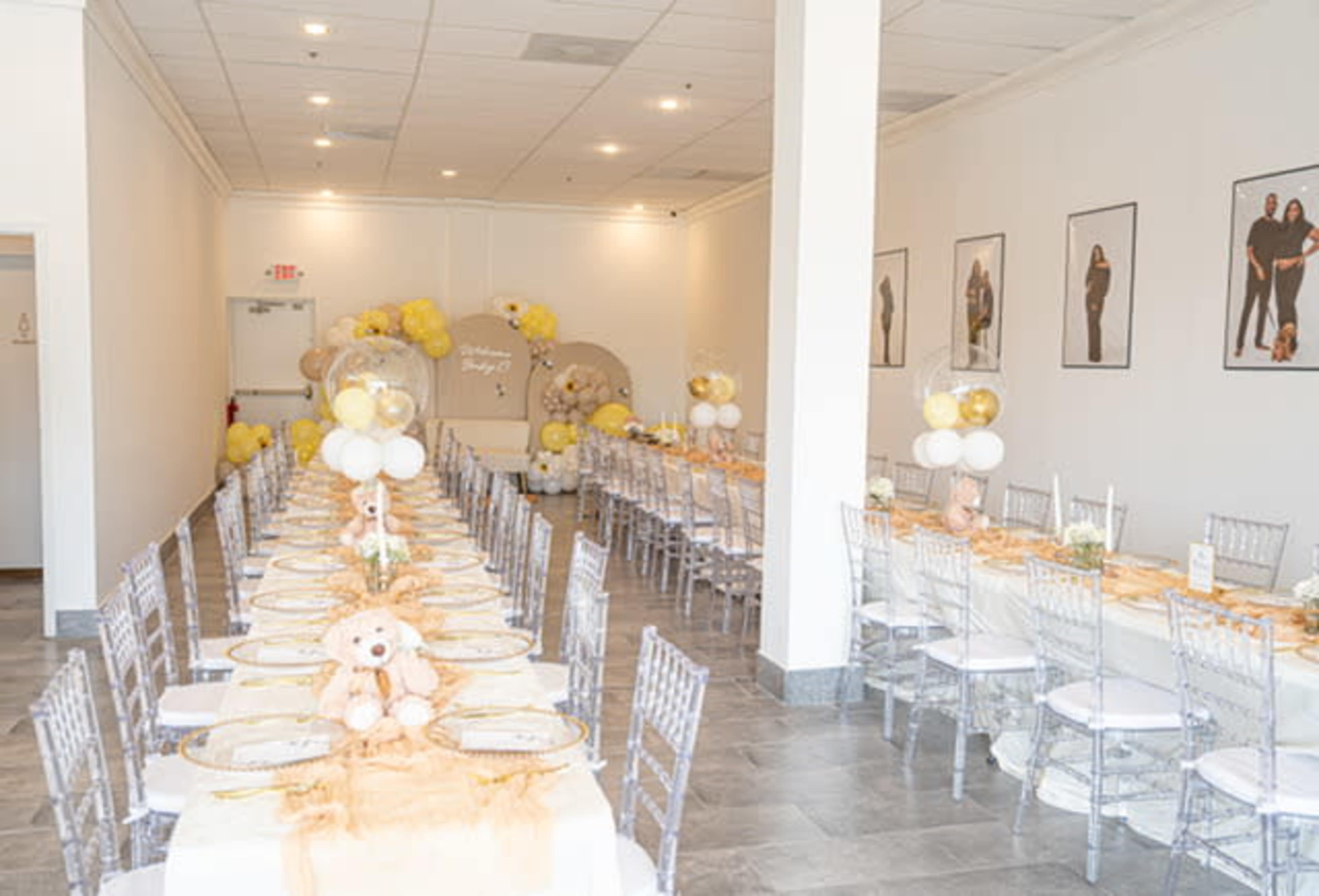 The image shows a banquet hall set with long tables adorned with white and gold decorations, featuring balloons and a teddy bear centerpiece.